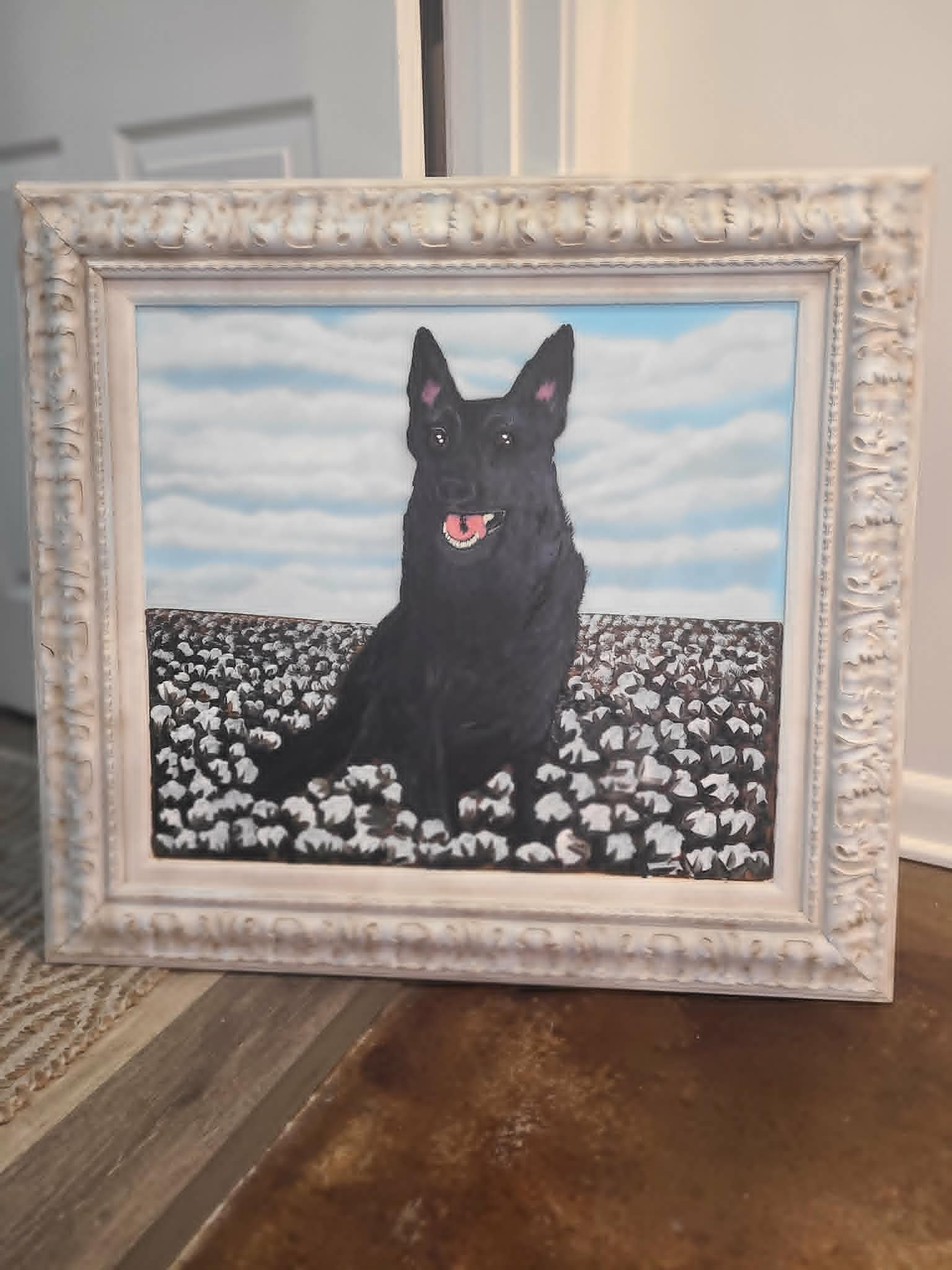Framed memorial portrait of a black German Shepherd sitting in a cotton field beneath a partly cloudy blue sky