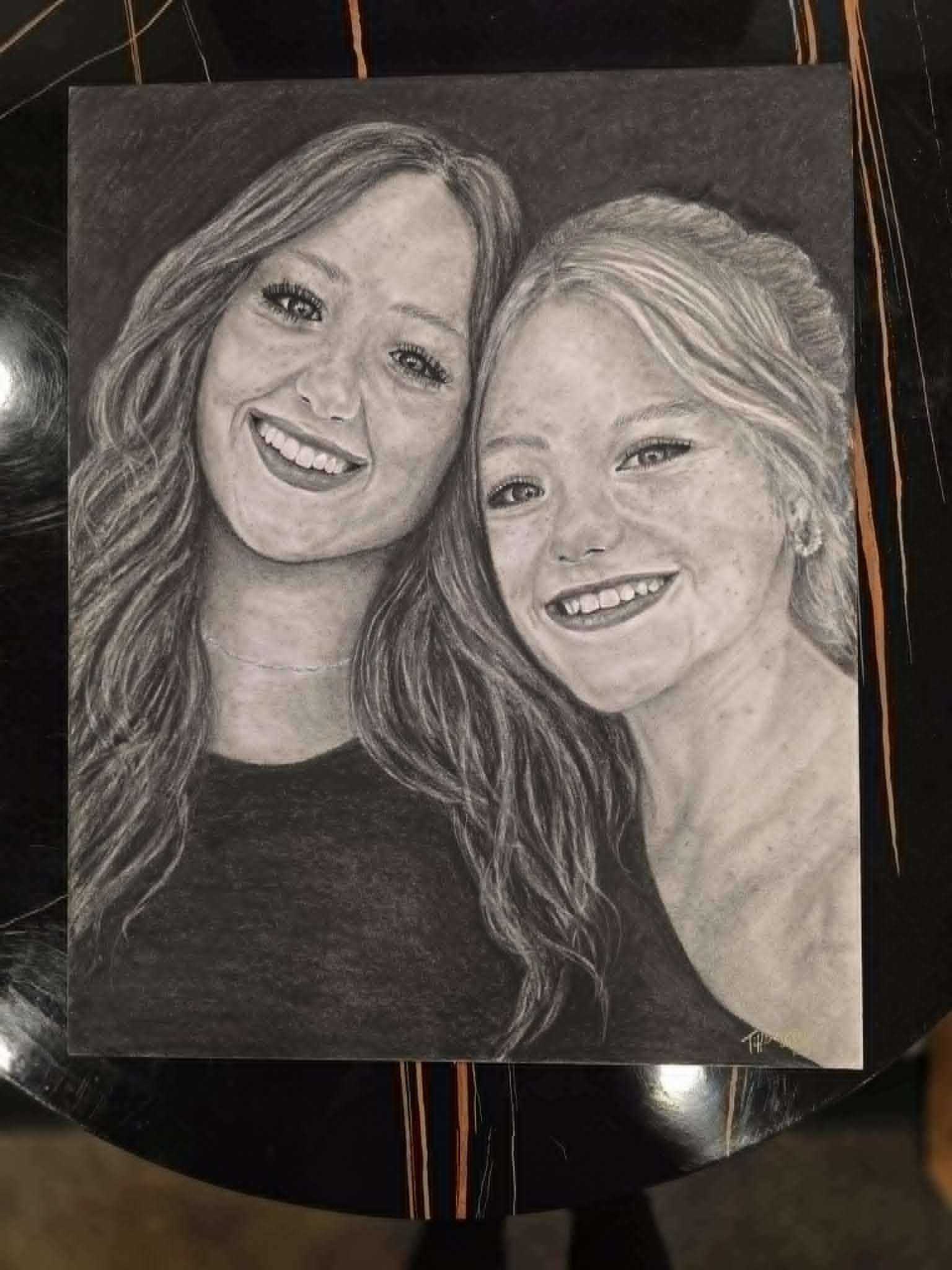 Detailed graphite portrait of two smiling young women side by side, displayed on a black marble table