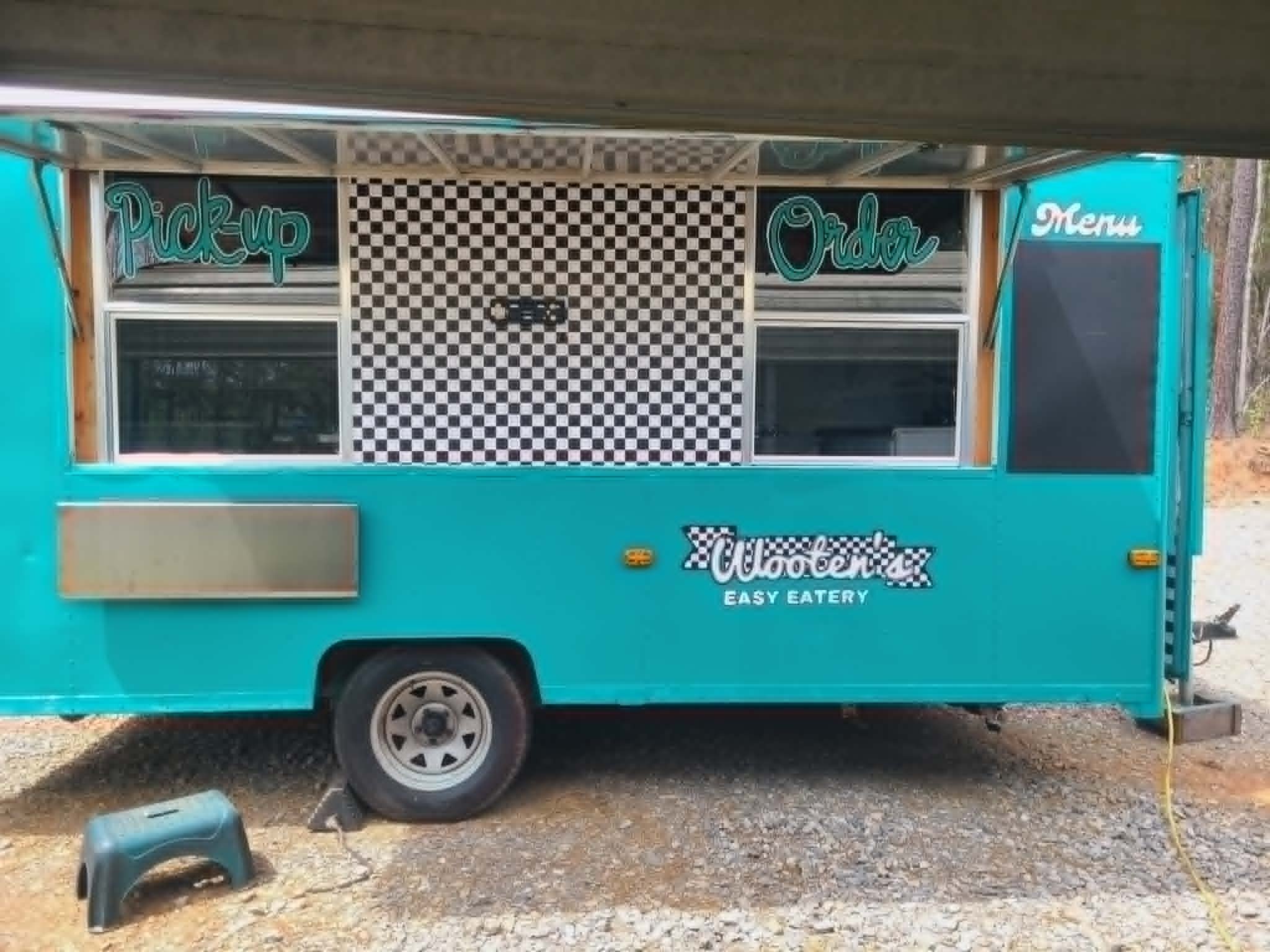 Side view of Wooten's Easy Eatery teal food trailer with hand-painted retro 'Pick-up' and 'Order' script window signs, black-and-white checkered panel, and Wooten's checkered-flag logo on the side