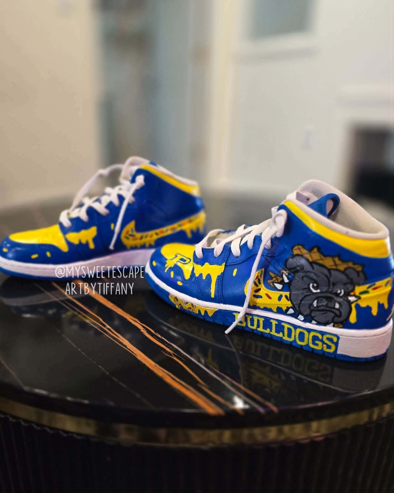 Hand-painted Piedmont Bulldogs Air Jordan 1 high-top sneakers in royal blue and yellow with the bulldog mascot, a dripping paint effect, and 'Bulldogs' team lettering on the heel