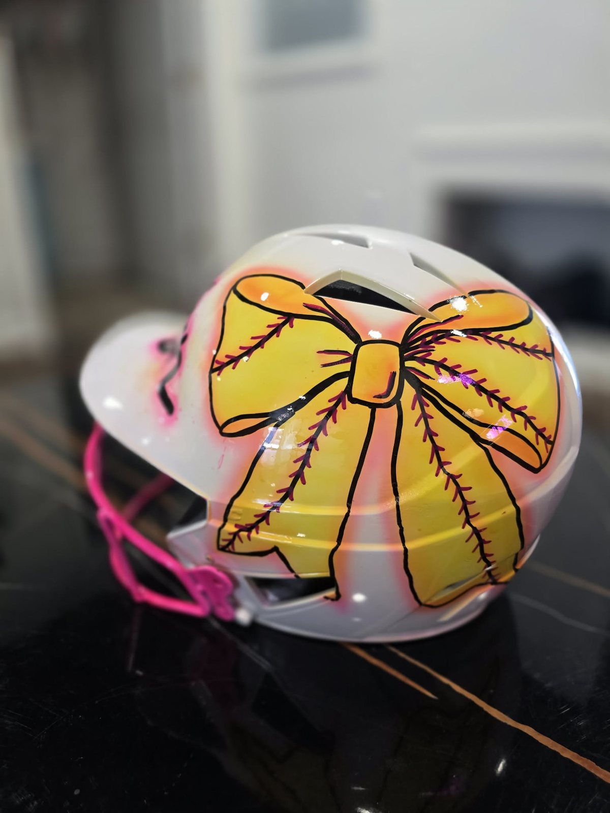Hand-painted white softball batting helmet with a large yellow softball-stitched bow in an airbrushed pink glow and a pink face guard