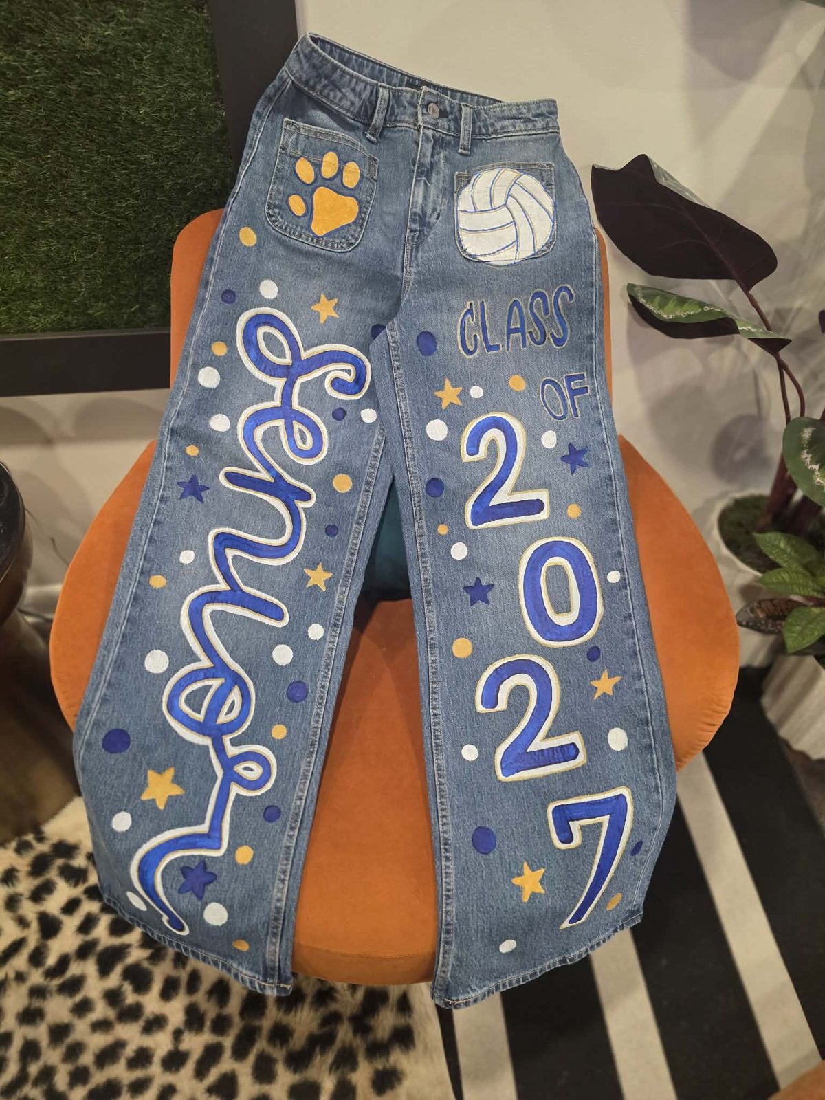 Hand-painted senior jeans in royal blue and gold with 'Senior' script down one leg, 'Class of 2027' down the other, a volleyball and paw print on the pockets, and scattered stars and dots