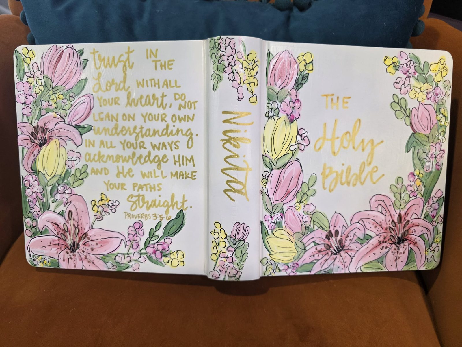 Hand-painted Bible cover for Nikita with pink lilies, tulips, and yellow blossoms framing 'The Holy Bible' in gold lettering, with Proverbs 3:5-6 hand-lettered on the back