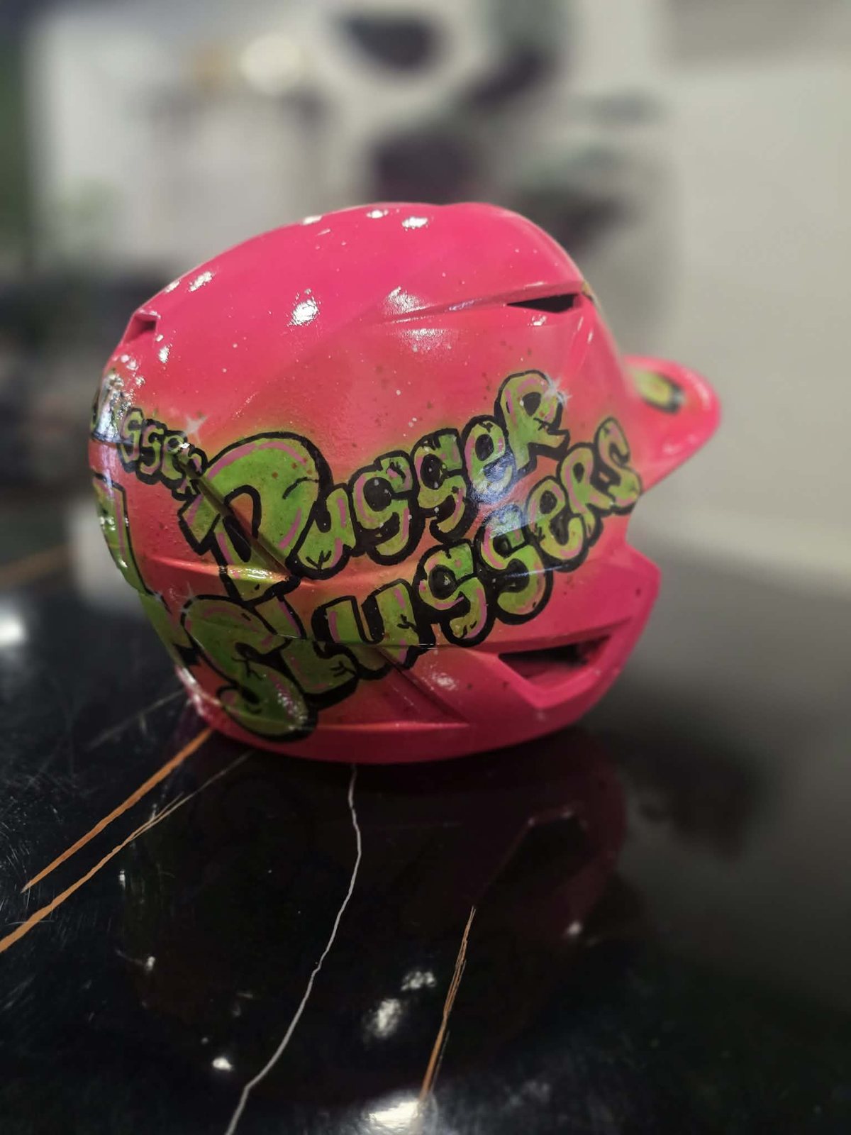 Hand-painted hot pink softball batting helmet with 'Pugger Sluggers' team name in lime green graffiti lettering and white paint speckles