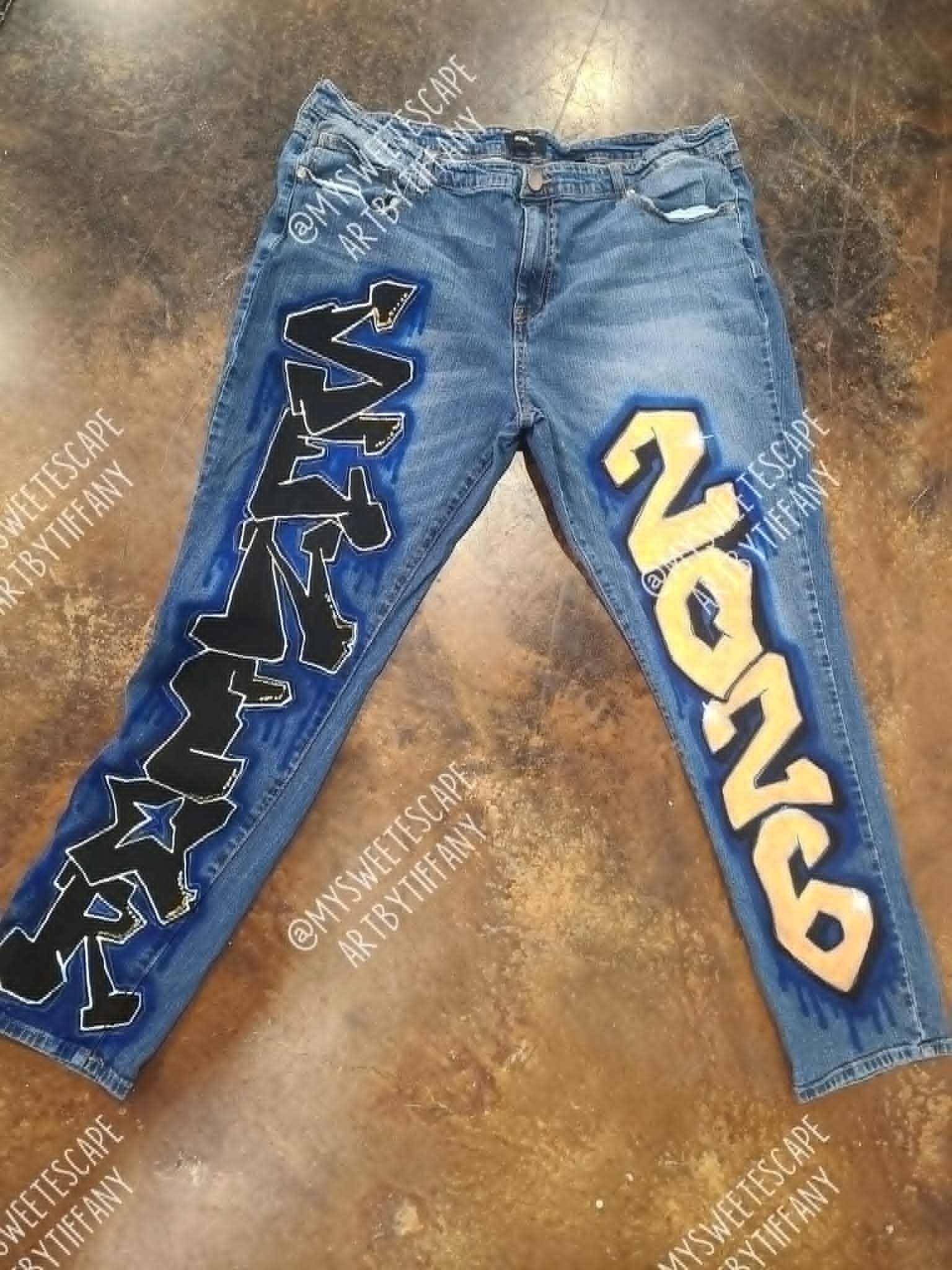Custom hand-painted blue jeans with bold black and cream graffiti-style lettering down each leg