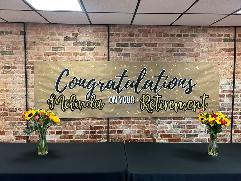 Hand-painted retirement banner reading 'Congratulations Melinda on your Retirement' in black and gold script with confetti accents, displayed on a brick wall with sunflower bouquets