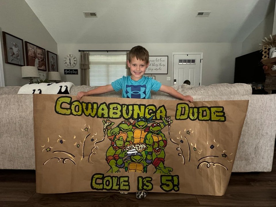 Hand-painted kraft paper birthday banner reading 'Cowabunga Dude, Cole is 5' featuring all four Teenage Mutant Ninja Turtles, held up by a smiling young boy