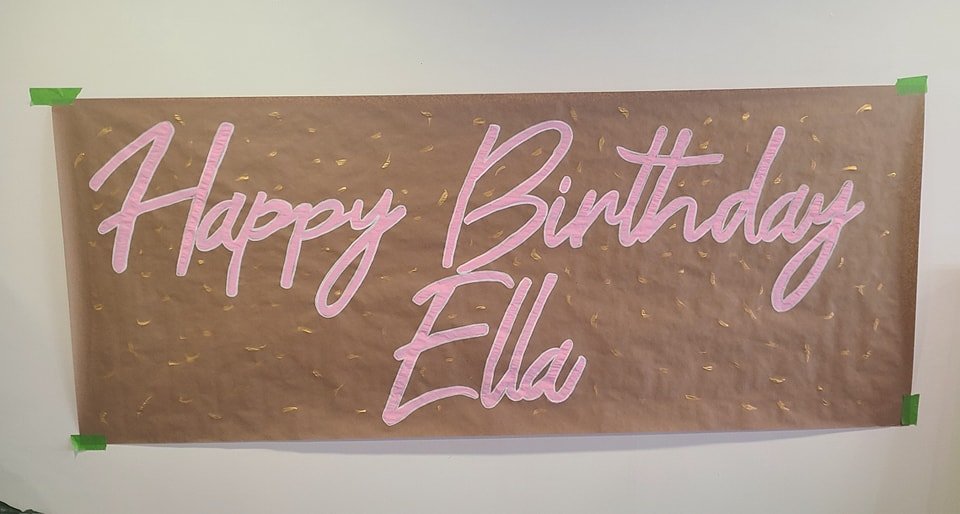 Hand-painted kraft paper birthday banner reading 'Happy Birthday Ella' in flowing pink script with gold confetti accents