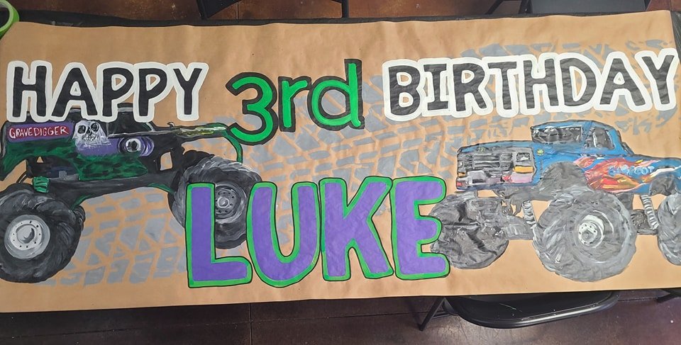 Hand-painted monster truck birthday banner reading 'Happy 3rd Birthday Luke' with Grave Digger and Bigfoot trucks on tire-tracked background