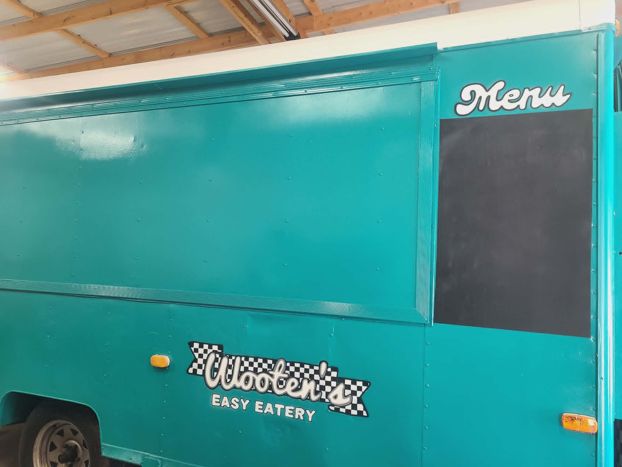 Side view of a teal food trailer with hand-painted Wooten's Easy Eatery checkered-flag logo and retro 'Menu' chalkboard panel