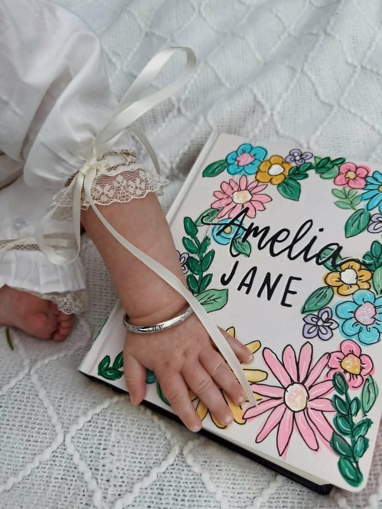 Hand-painted Bible cover for Amelia Jane with colorful floral wreath, photographed beside a baby in a christening gown