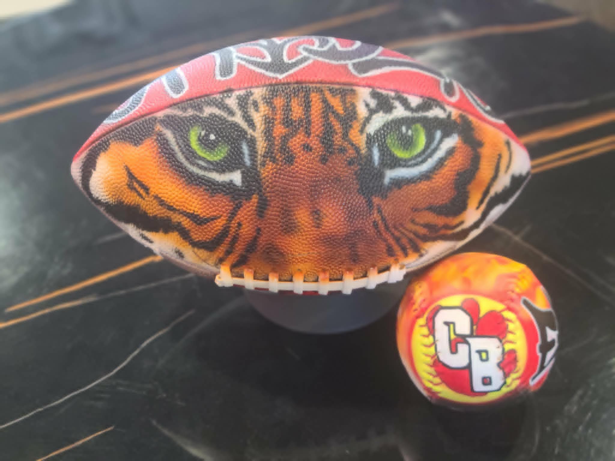 Hand-painted football with realistic green-eyed tiger face and red graffiti lettering, displayed beside an airbrushed yellow softball with red CB lettering and flames