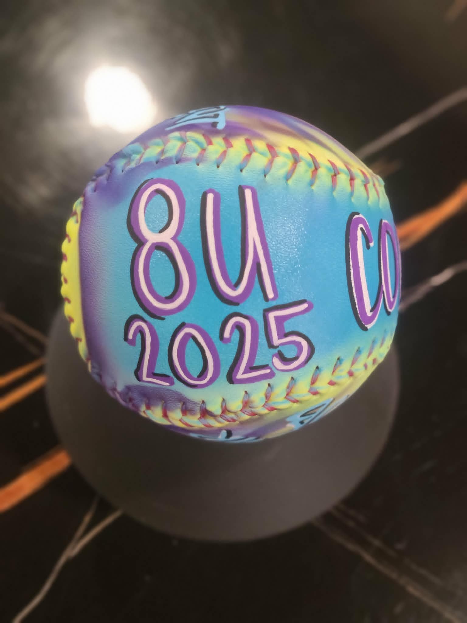 Airbrushed pastel softball with blue, purple, and yellow tie-dye fade and '8U 2025' team lettering in purple bubble font