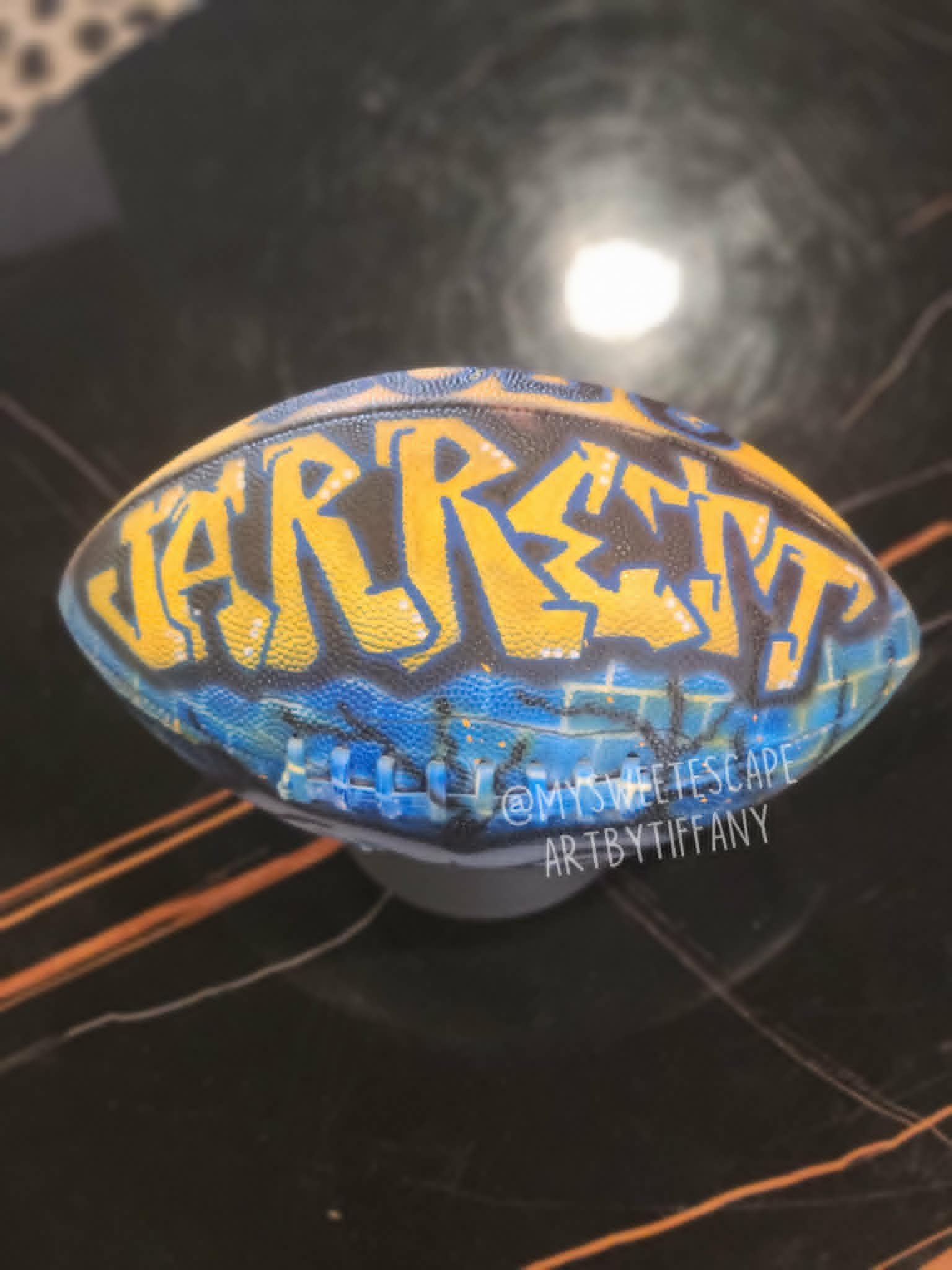 Hand-painted football with 'Jarrett' in bold yellow graffiti lettering and blue brick wall background