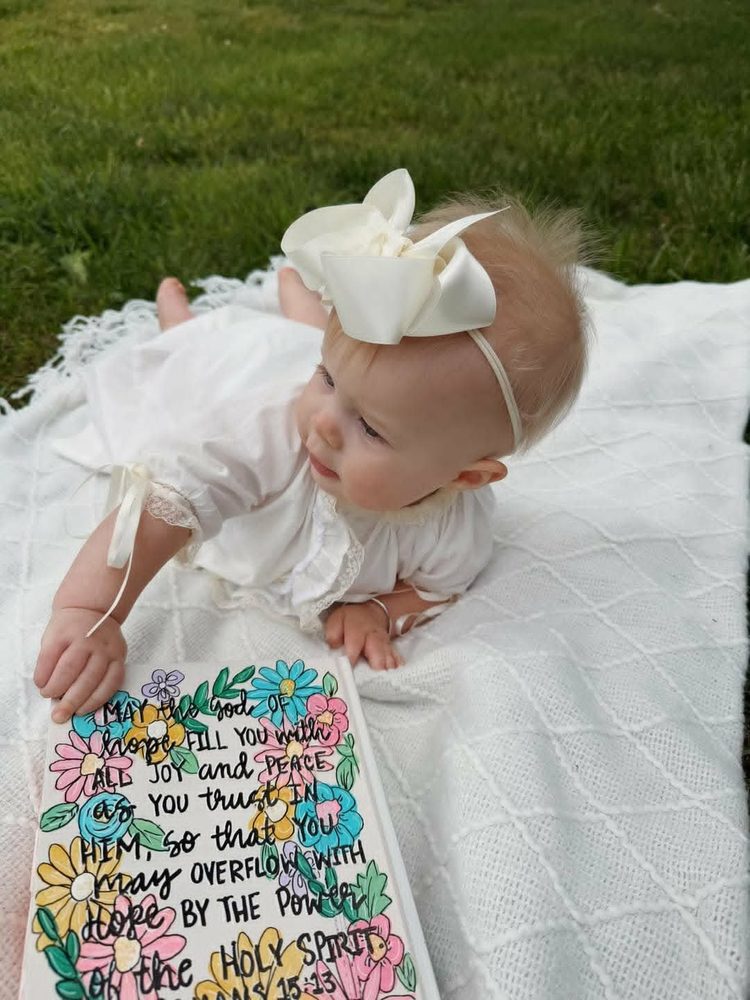 Hand-painted Bible cover with Romans 15:13 verse and pastel florals, beside a baby girl in a white bow
