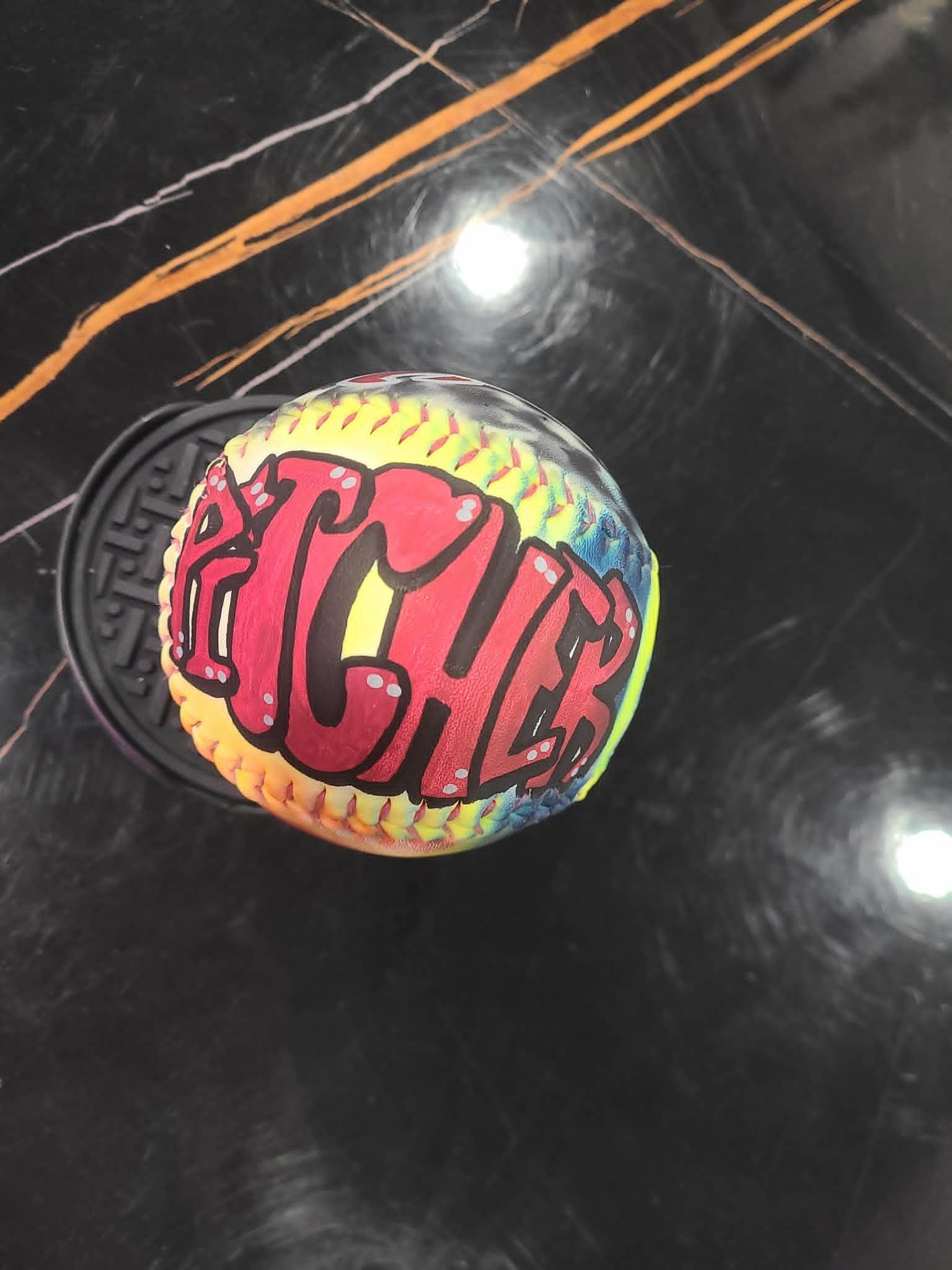 Hand-painted softball with bold red graffiti 'Pitcher' lettering on yellow and blue tie-dye background