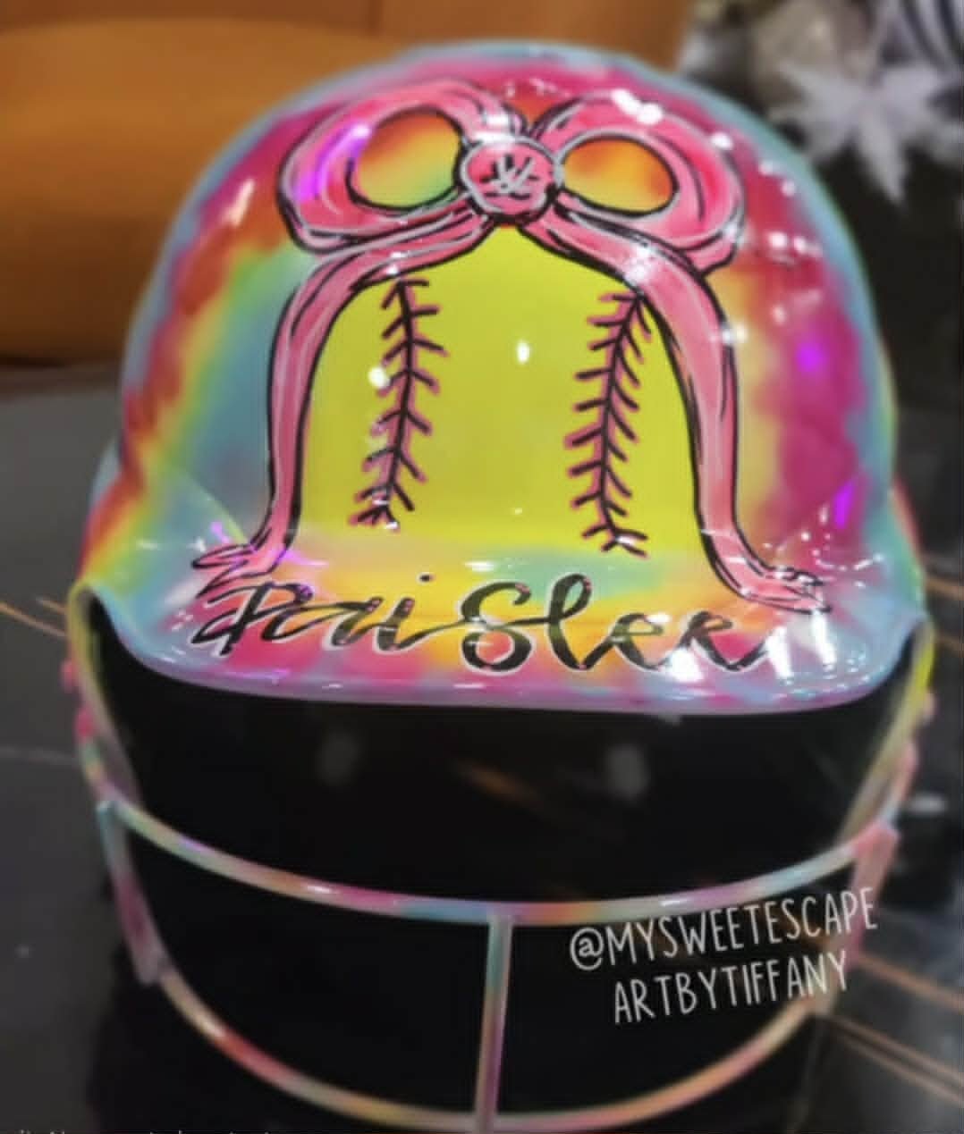 Custom airbrushed catcher's helmet for Paisley with pastel rainbow tie-dye, hand-painted softball wrapped in a pink bow, and personalized script lettering