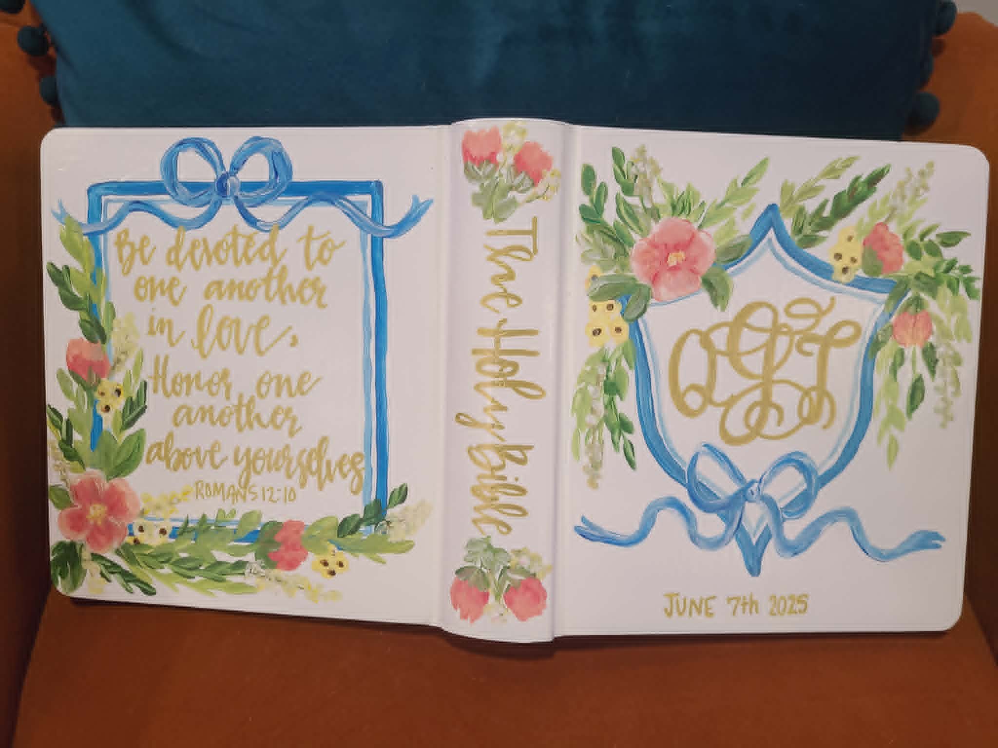 Hand-painted wedding Bible with gold monogram crest, blue ribbon bows, pink florals, Romans 12:10 verse, and June 7th 2025 wedding date