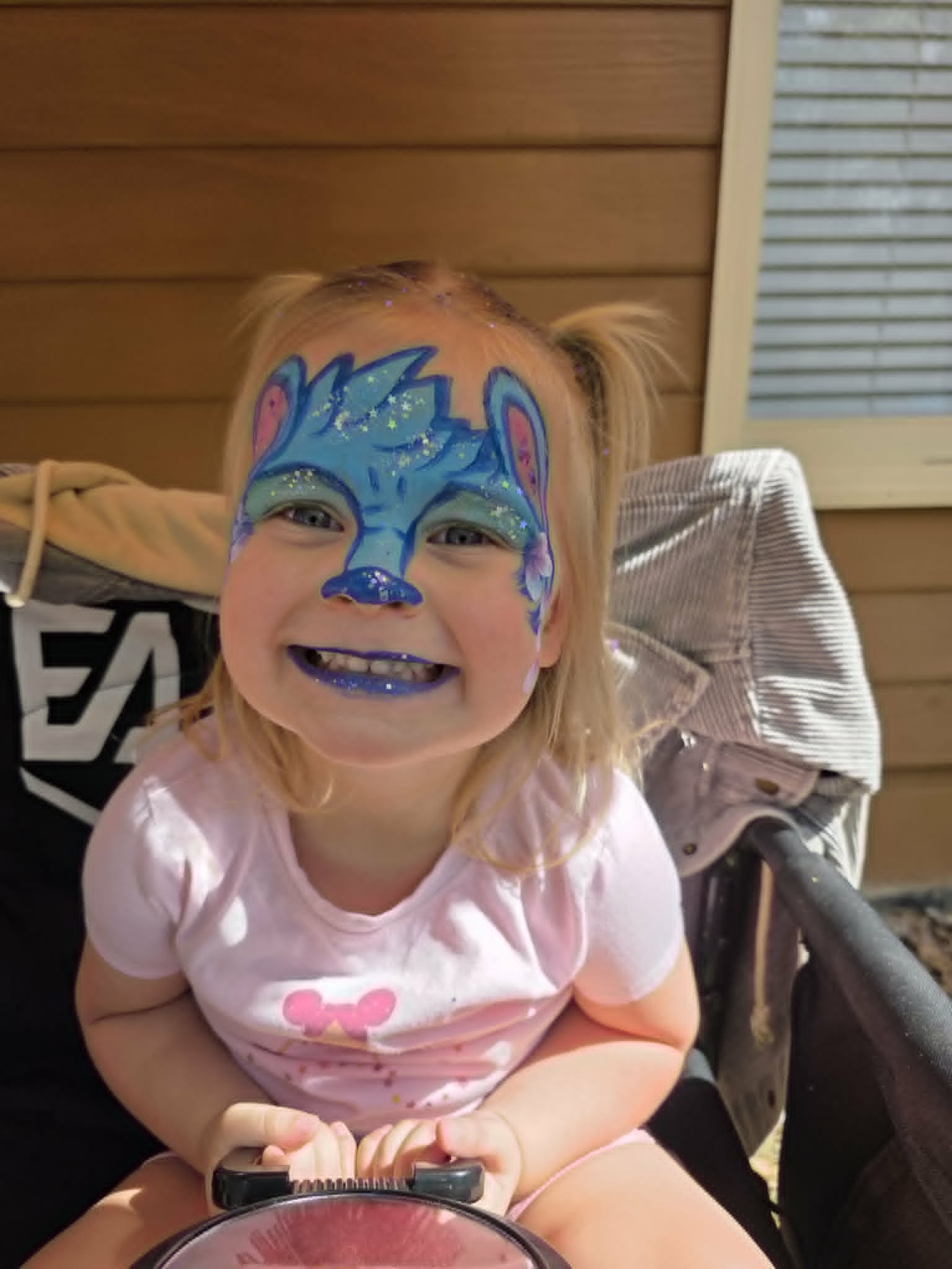 Sparkly blue wolf or fox face paint with pink ears and glitter accents on a smiling toddler girl