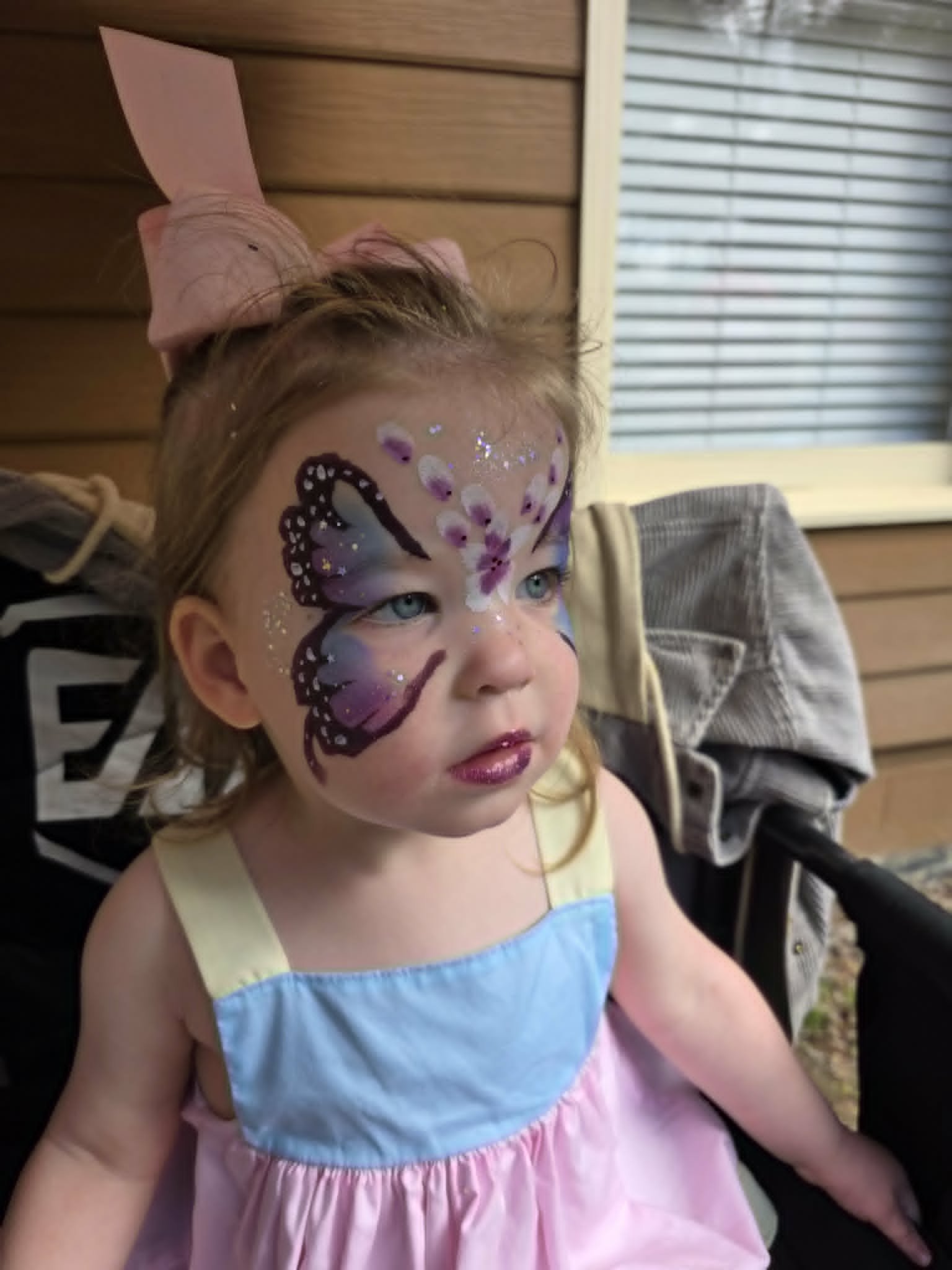 Elegant purple butterfly face paint with glitter accents and matching glossy lips on a young girl with a pink bow