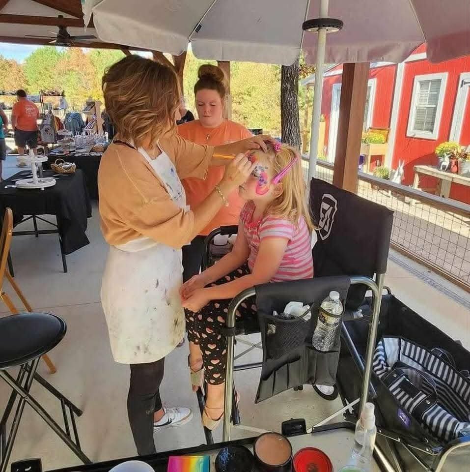 Tiffany painting a young girl's face under an outdoor umbrella tent at a fall festival