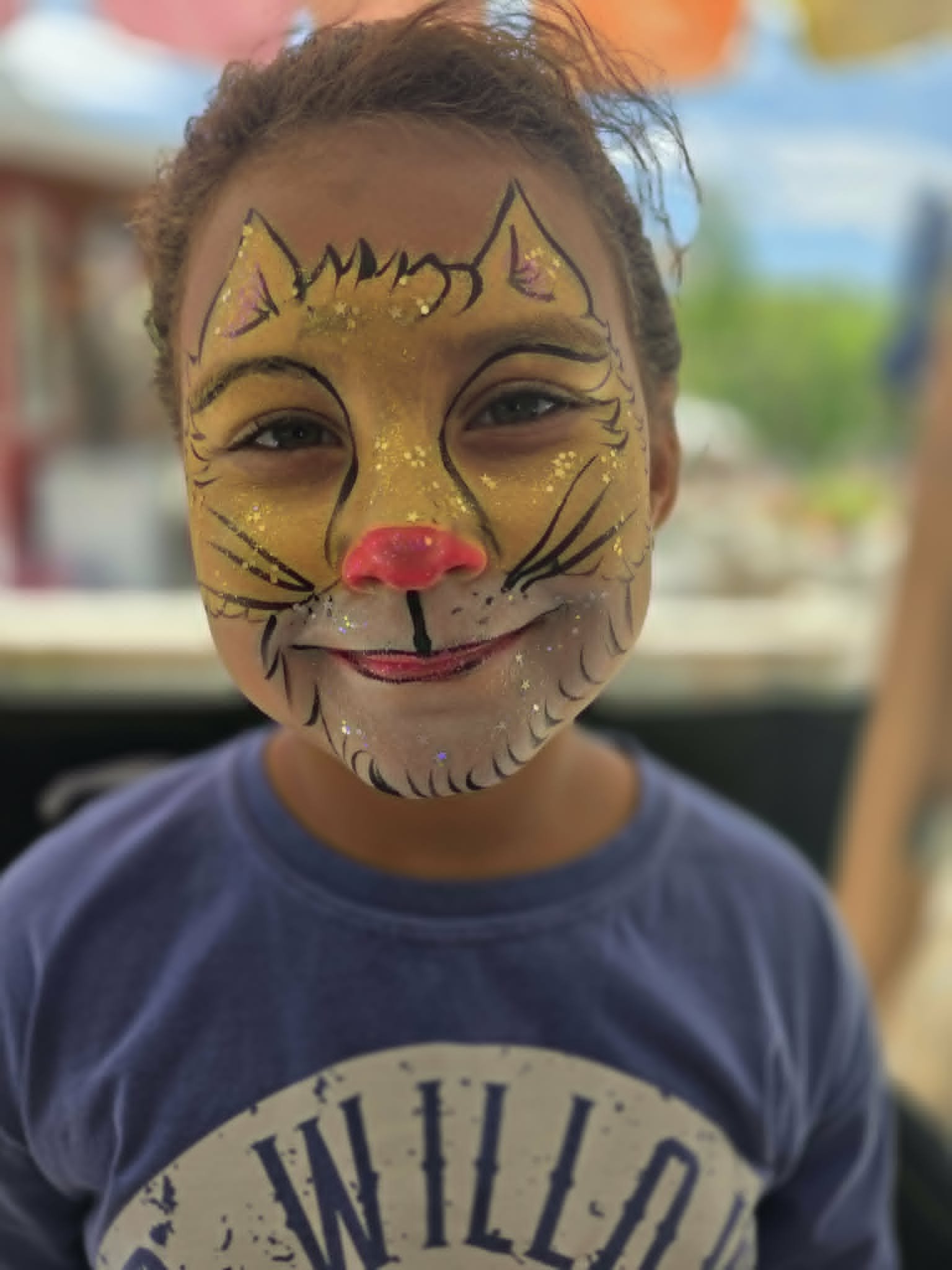 Glittery gold tiger face paint with pink nose and detailed whiskers on a young girl