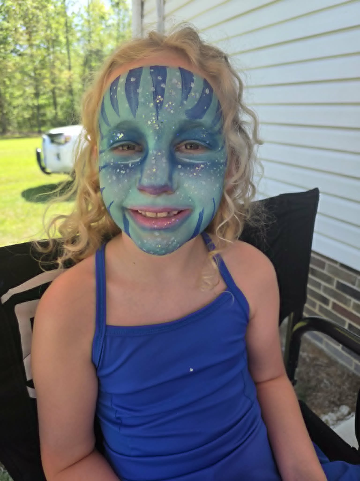Mystical blue and green Avatar-inspired full-face paint with stripes and gold glitter freckles on a young girl