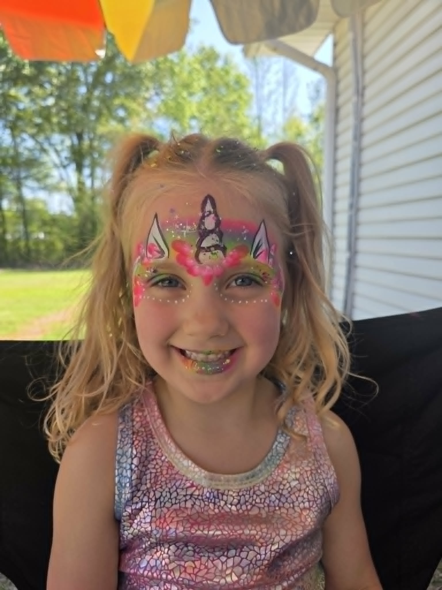 Rainbow unicorn face paint with glittery horn, ears, and pastel colors on a smiling young girl with pigtails