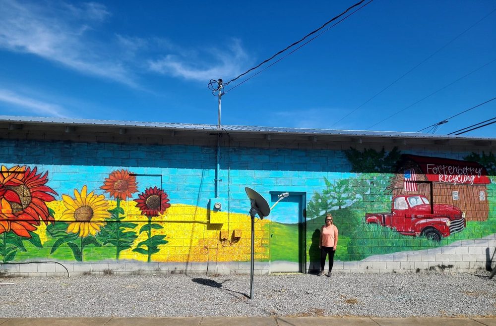 Wide outdoor mural of sunflowers, blue sky, and a red farm truck for Fortenberry Recycling, with Tiffany standing in front