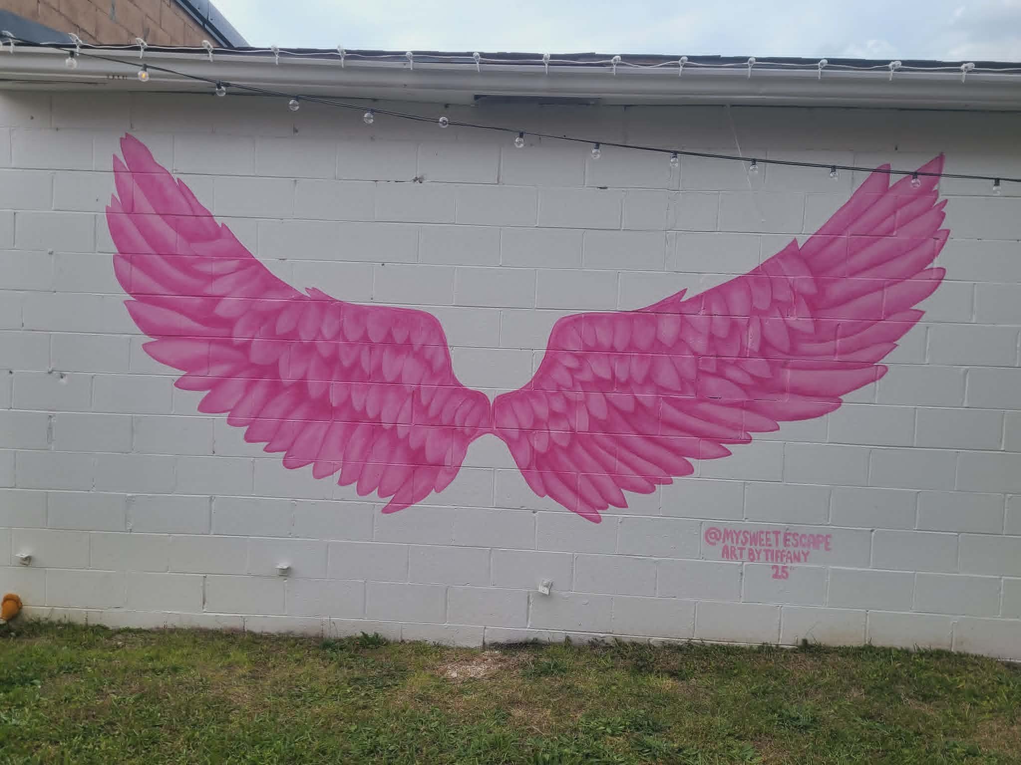Large pink angel-wing photo-op mural on a white cinderblock wall with cafe string lights overhead