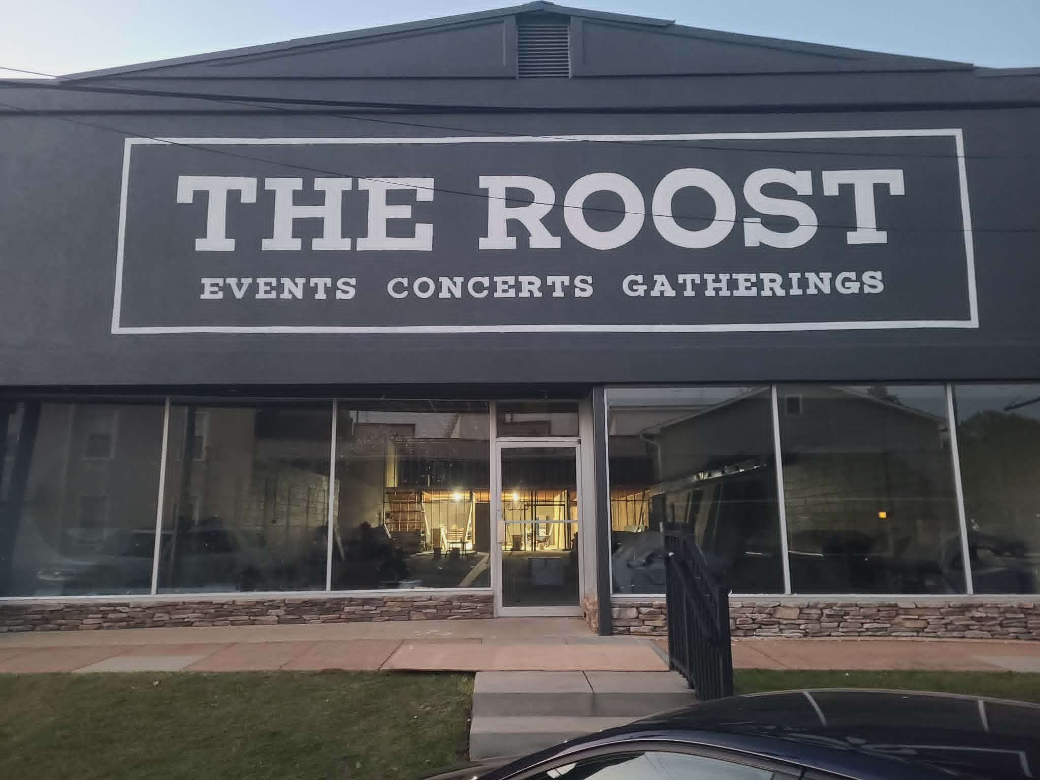 Hand-painted 'The Roost — Events Concerts Gatherings' storefront sign in clean white block lettering on a dark gray facade