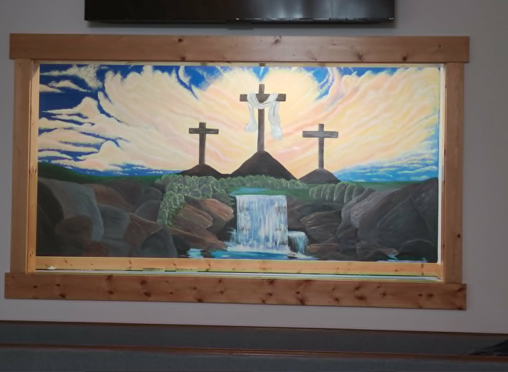 Framed indoor mural of three crosses on a hillside with a draped white cloth, glowing sunset clouds, and a waterfall flowing through rocky terrain
