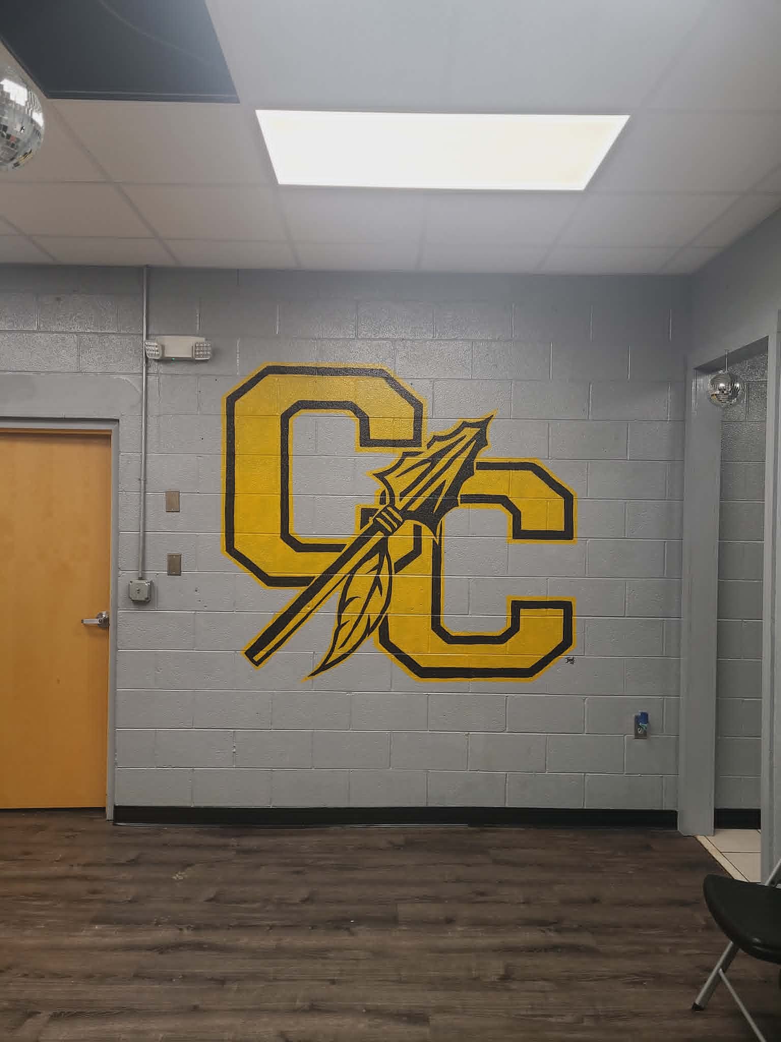 Cherokee County 'CC' varsity logo mural with a tomahawk and feathers, painted in bold yellow and black on a school cinderblock wall