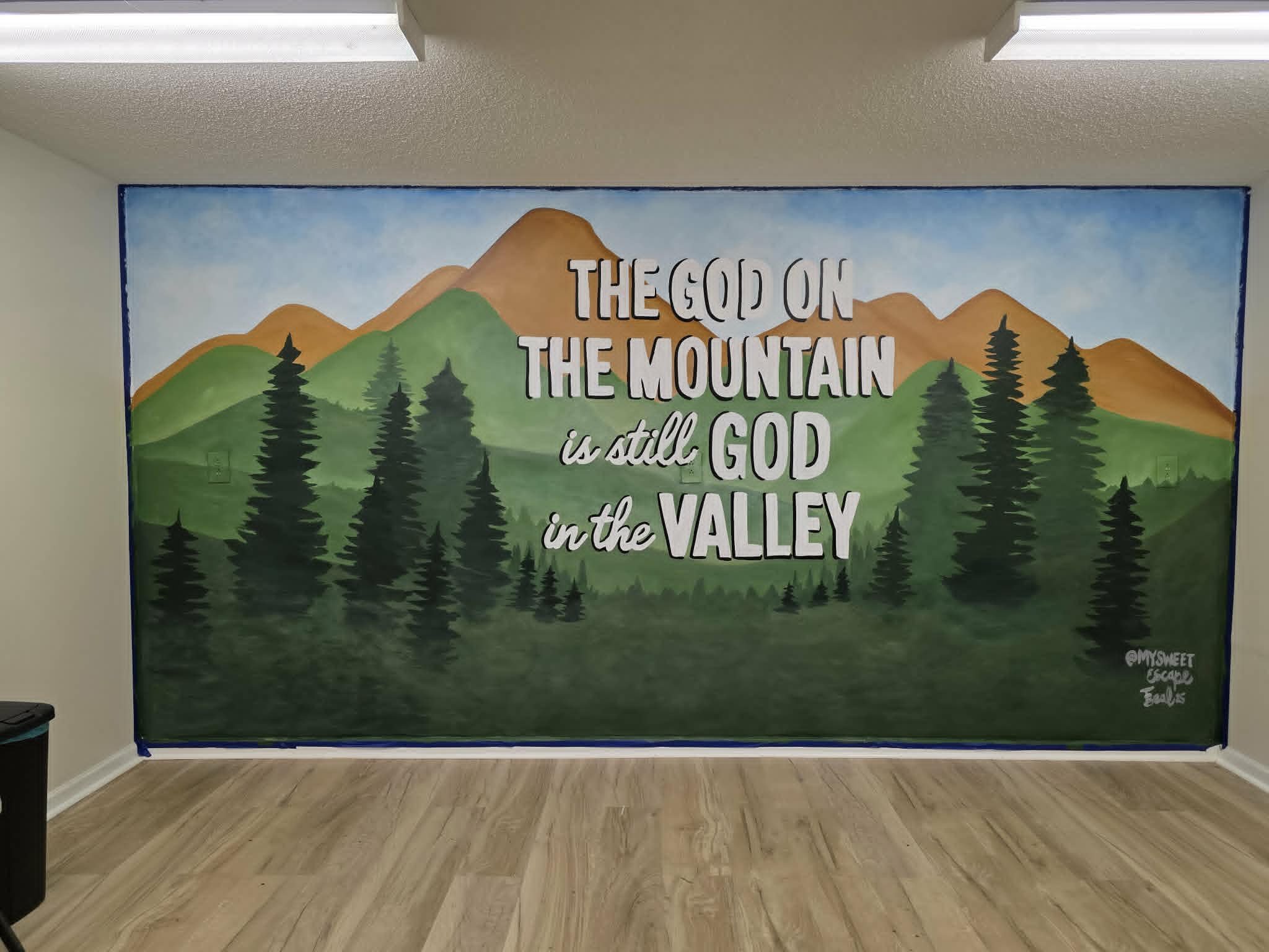 Mountain landscape mural with rolling green hills, tall pine trees, and a scripture-inspired quote 'The God on the Mountain is still God in the Valley' lettered in white