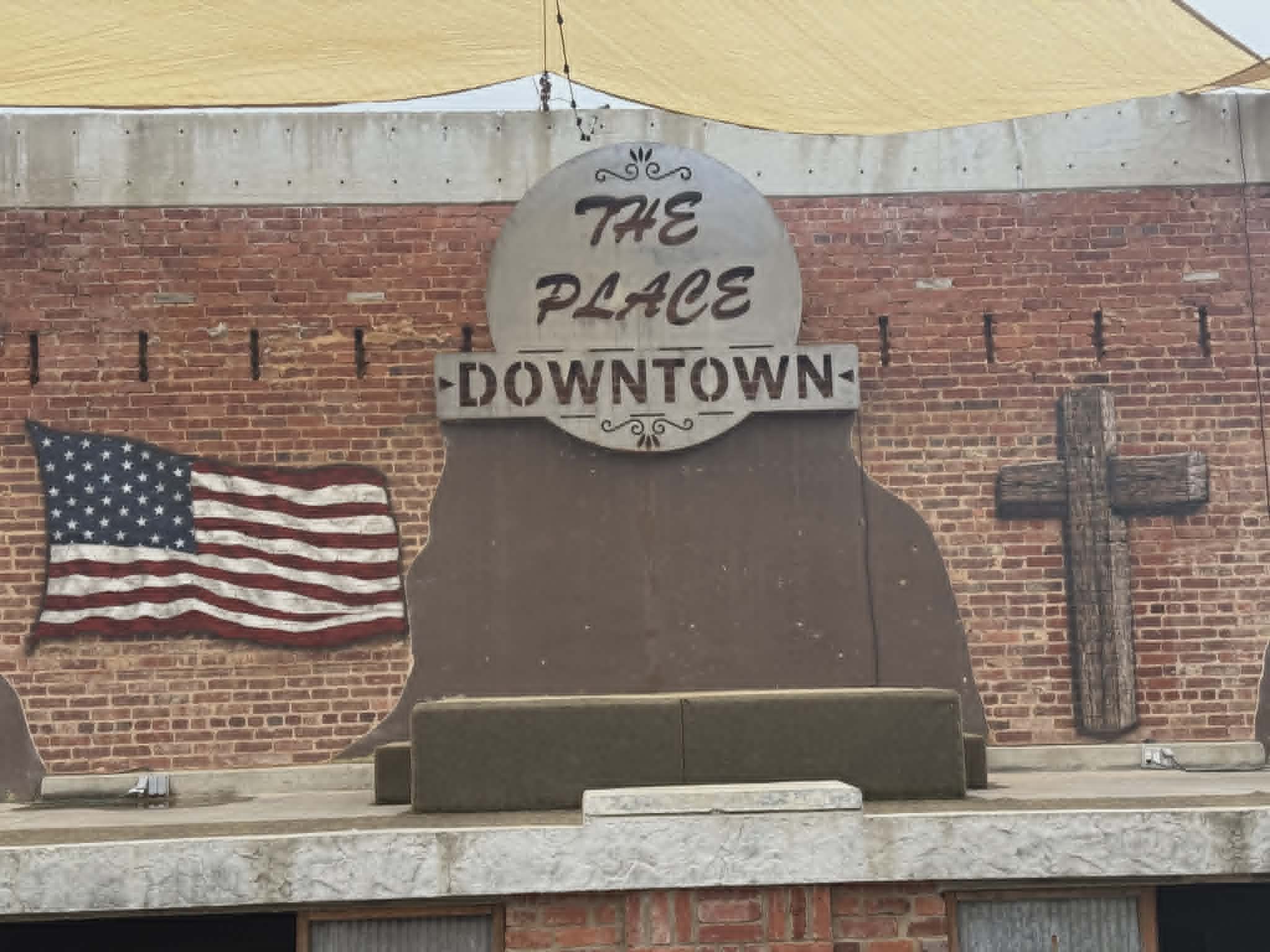 The Place Downtown exterior brick wall mural with a hand-painted American flag, a rustic wooden cross, and a circular 'The Place Downtown' sign mounted above a stage