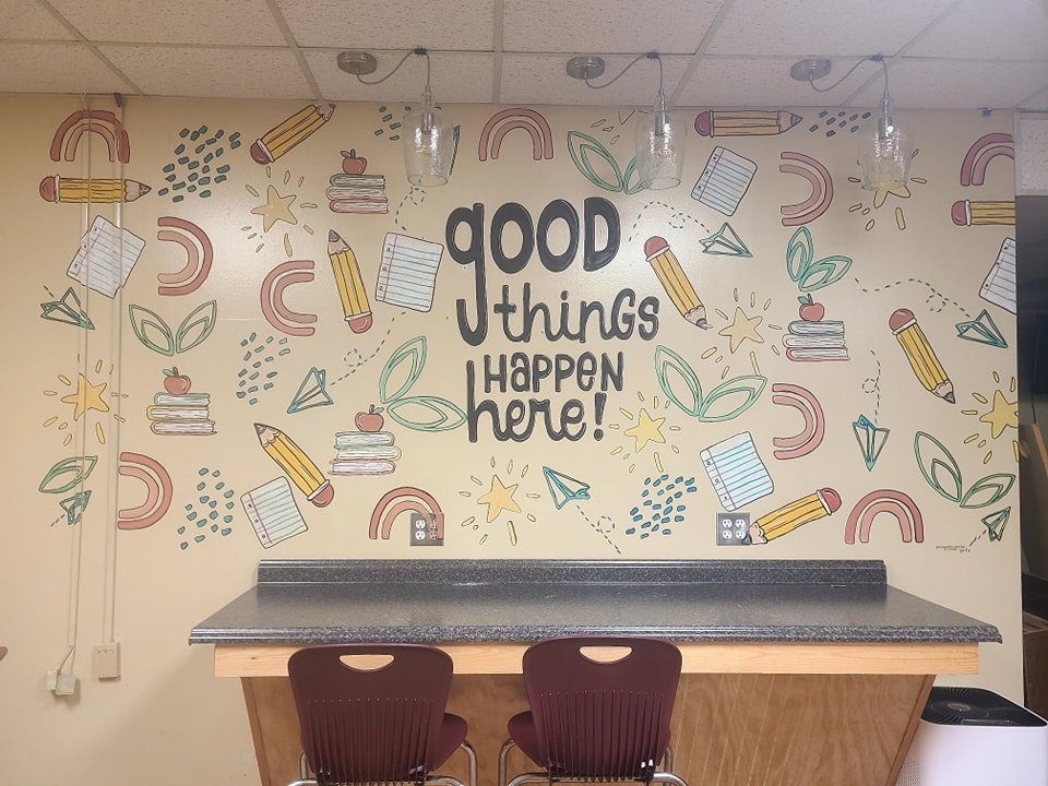 School classroom mural reading 'good things happen here' surrounded by hand-drawn pencils, books, rainbows, and stars