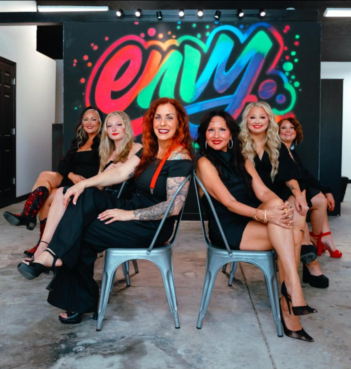 Envy Salon team posing in black dresses on metal chairs in front of the colorful Envy graffiti mural