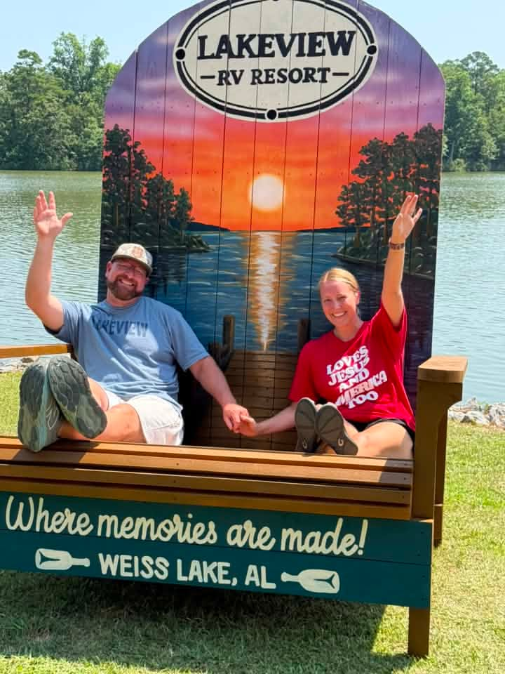 A couple seated in the Lakeview RV Resort giant painted Adirondack chair at Weiss Lake, AL, with its lake sunset mural and 'Where memories are made' lettering