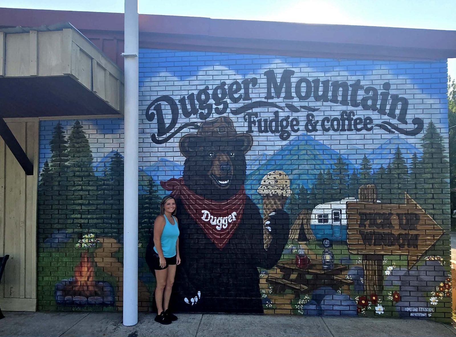Dugger Mountain Fudge & Coffee exterior brick mural of a bear in a ranger hat and red bandana holding an ice cream cone, with pine trees, blue mountains, a campfire, a vintage camper and a wooden pick-up window sign