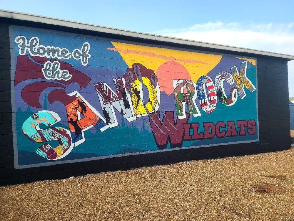 Home of the Sand Rock Wildcats postcard-style lettering mural with mountain and farm scenes inside each letter