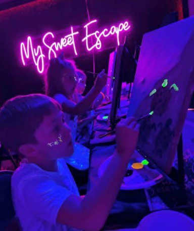Young boy painting at an easel under the My Sweet Escape neon sign with blacklight glow