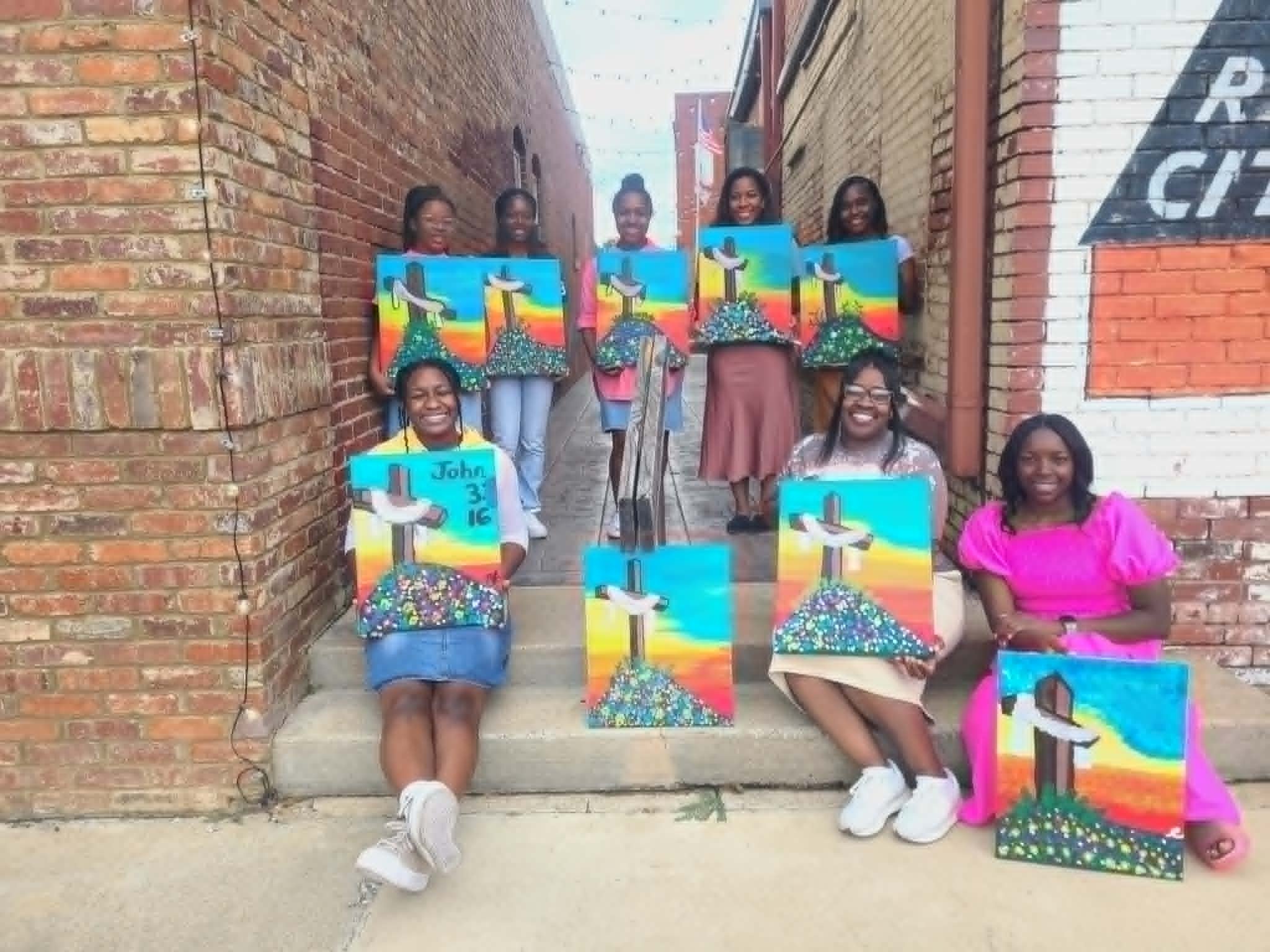 Church youth group posing in a downtown alley with their finished cross paintings featuring rainbow sunset skies and beds of wildflowers, with John 3:16 lettered on one canvas
