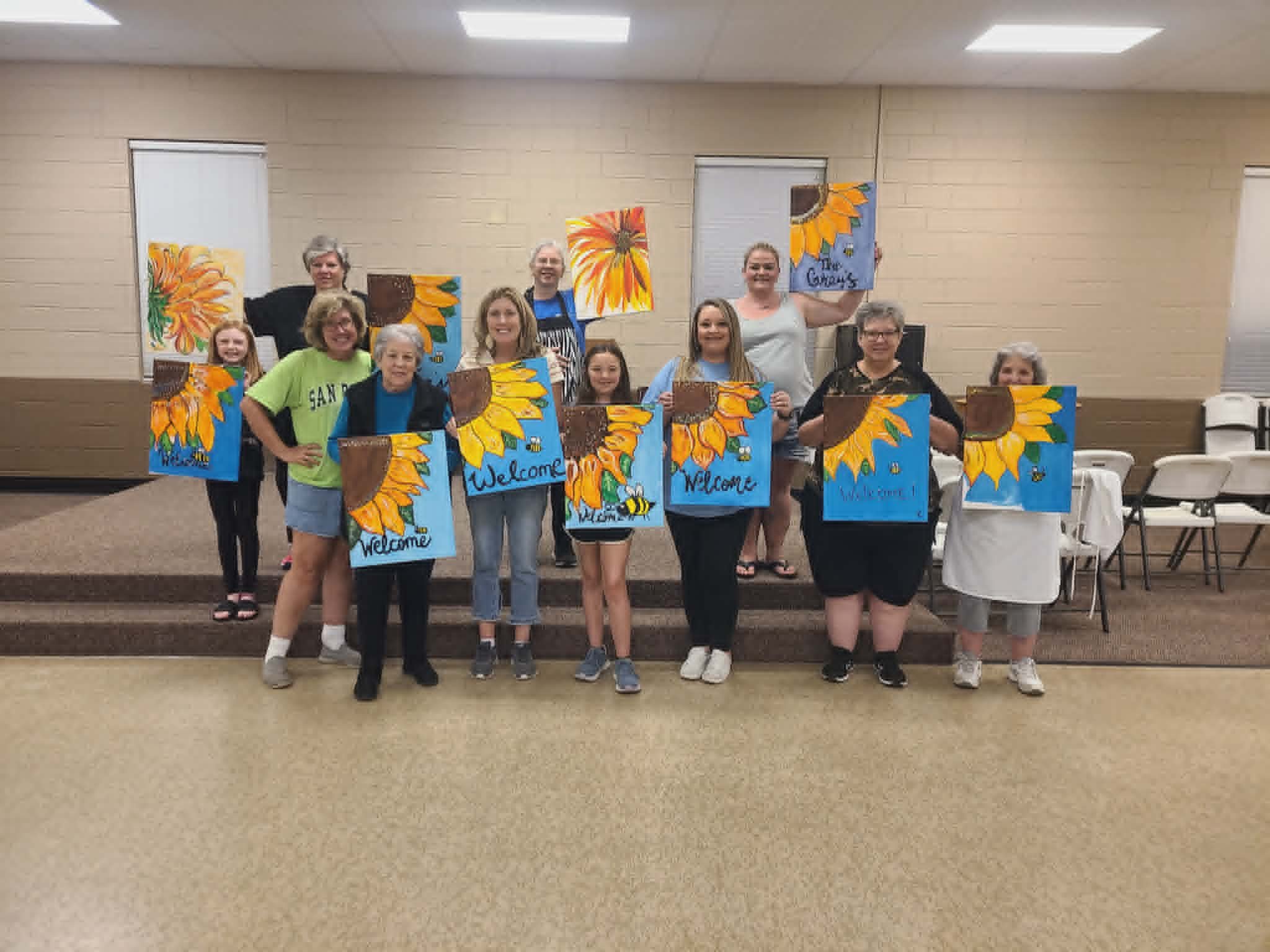 Multi-generational church group in a fellowship hall holding up their finished sunflower 'Welcome' paintings with bumblebee accents