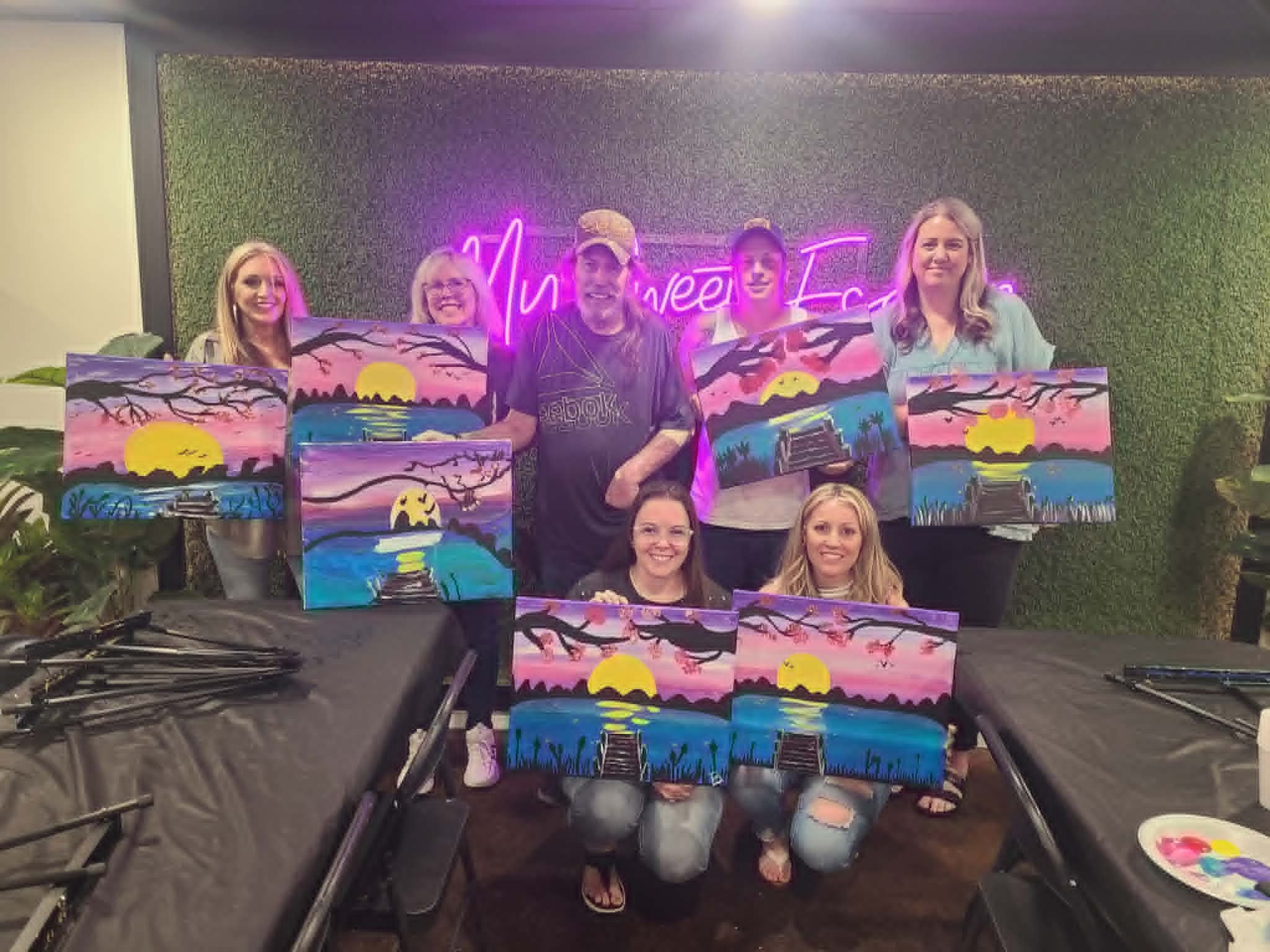 Adult paint party group in the My Sweet Escape studio holding up their finished cherry-blossom sunset and dock paintings under the neon sign