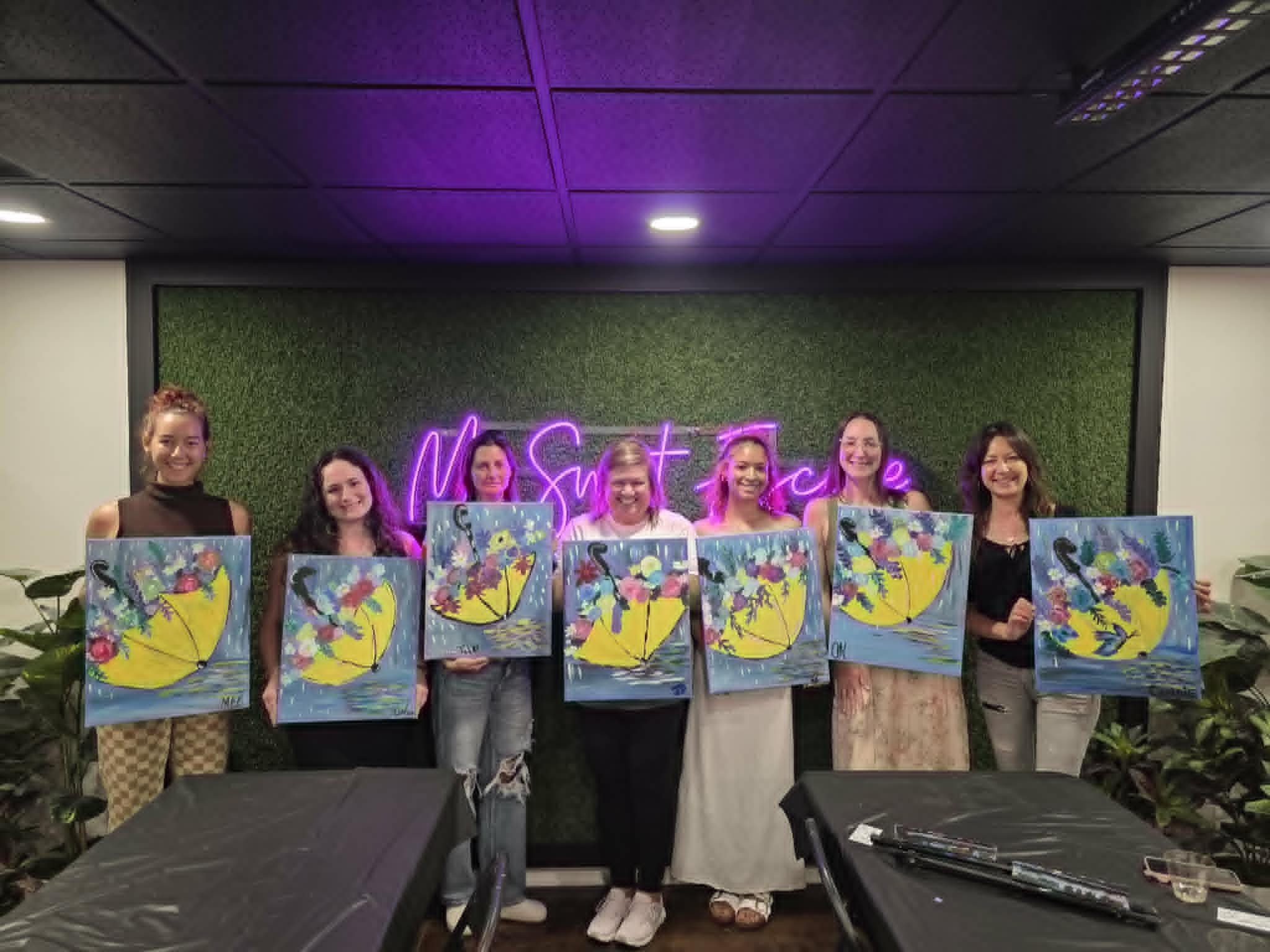 Group of women in the My Sweet Escape studio holding up their finished yellow umbrella with floral bouquet rainy-day paintings