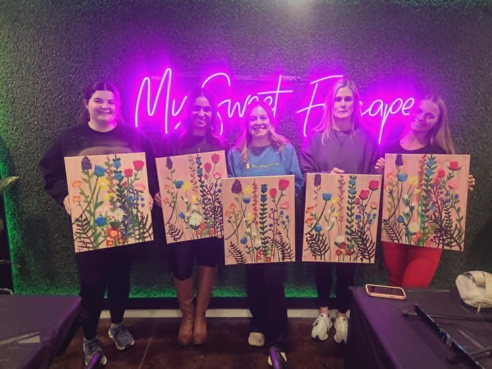 Group of women in the My Sweet Escape studio holding up their finished wildflower meadow paintings with vibrant blooms on a soft pink background