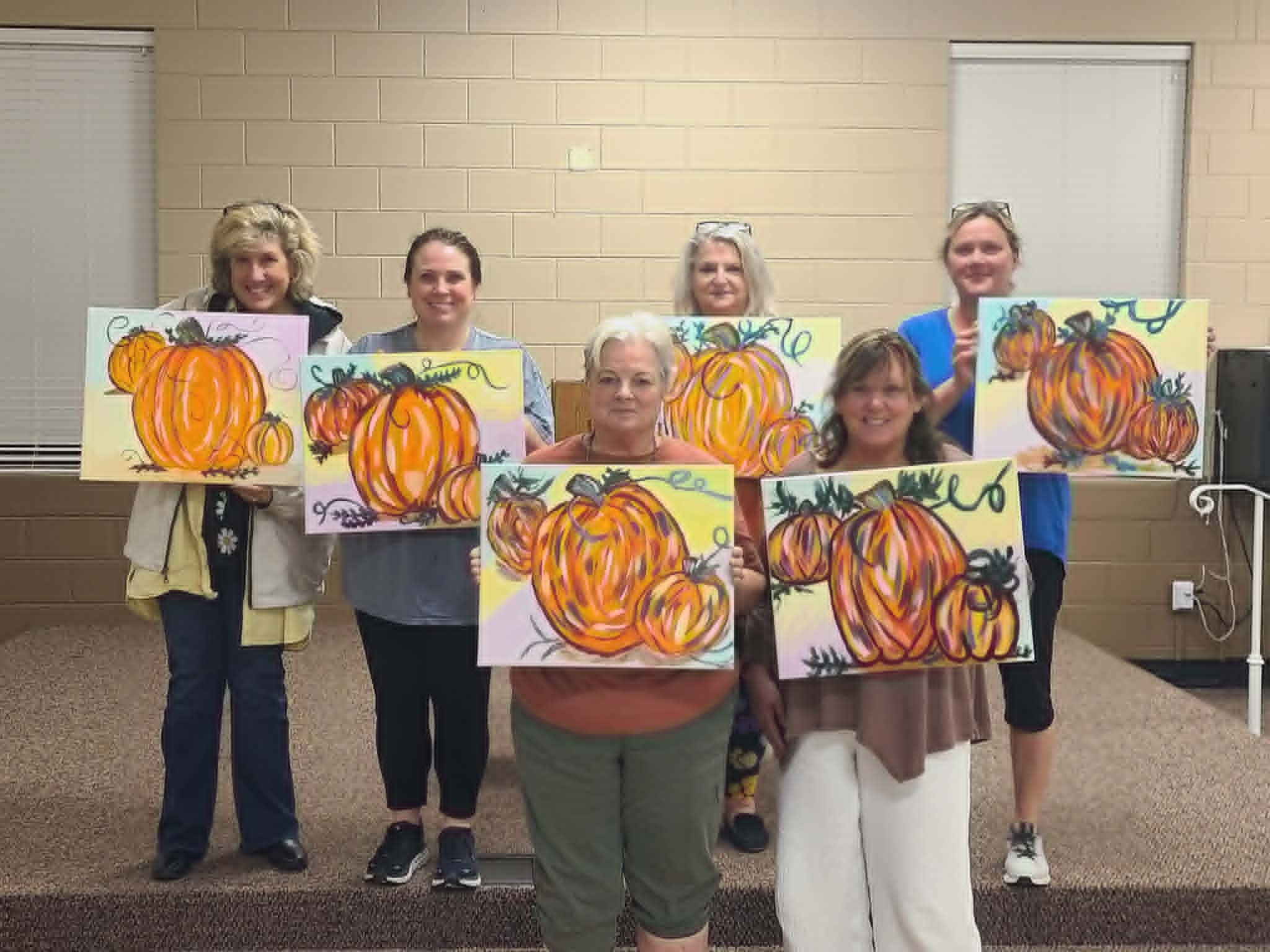 Group of women holding up their finished expressive pumpkin paintings in vibrant orange brushstrokes on pastel yellow and pink backgrounds
