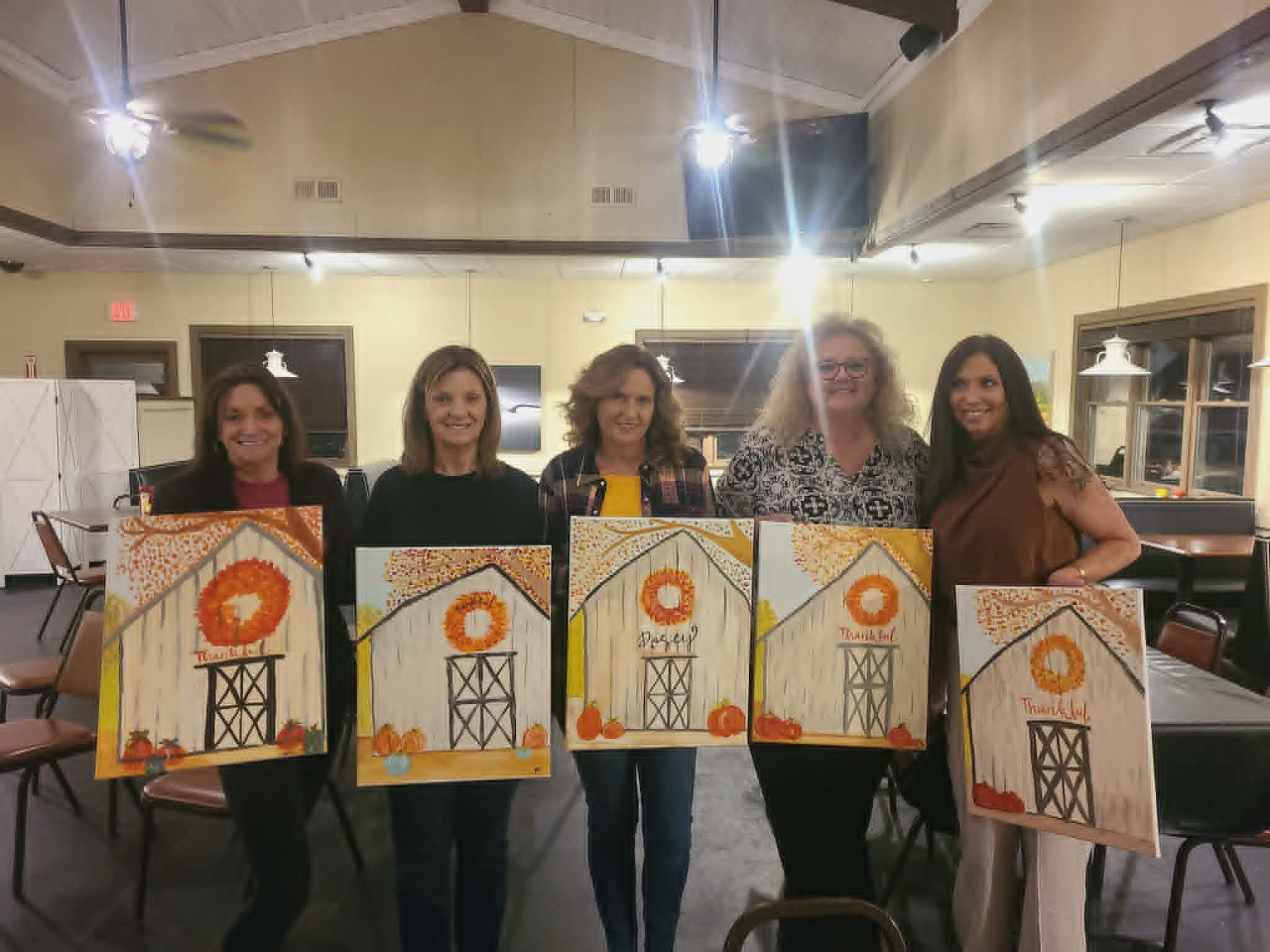 Group of women holding up their finished autumn barn paintings featuring a white barn with orange wreath and falling leaves