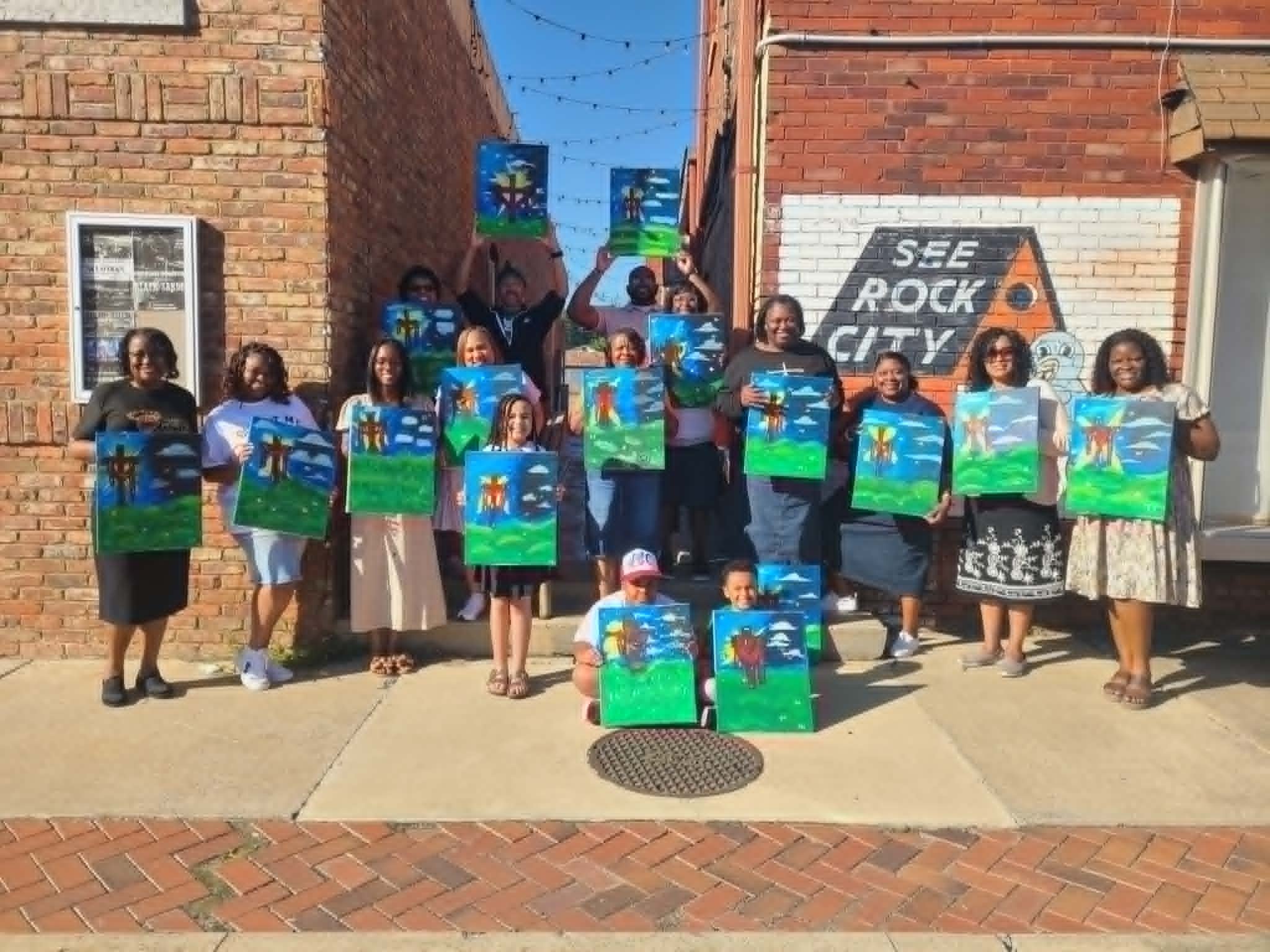 Outdoor downtown paint party group holding up their finished three-crosses-on-a-hill paintings with bright blue skies and green grass