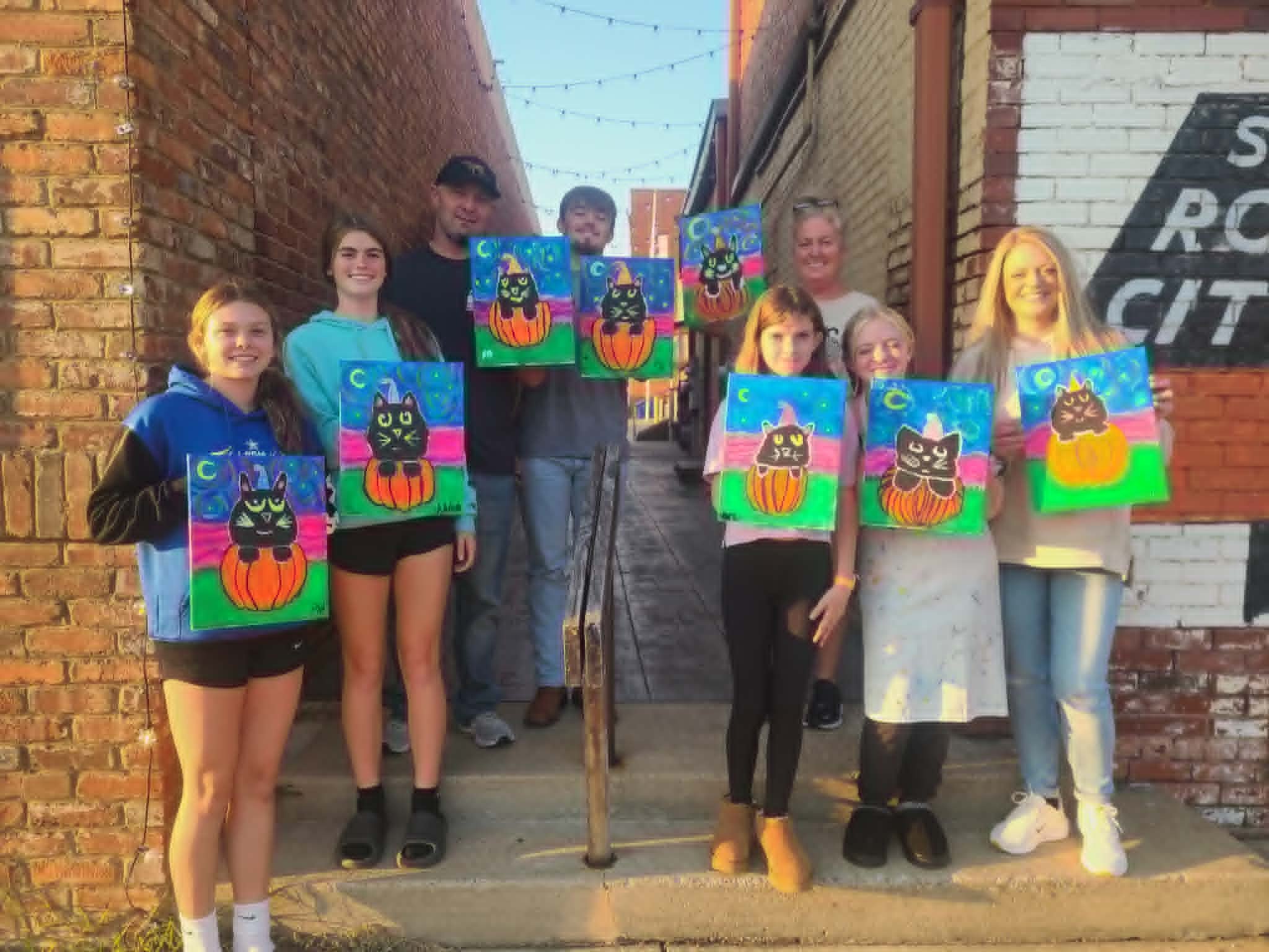 Halloween paint party group in a downtown alley holding up their finished black cat in a pumpkin paintings on starry purple and pink backgrounds